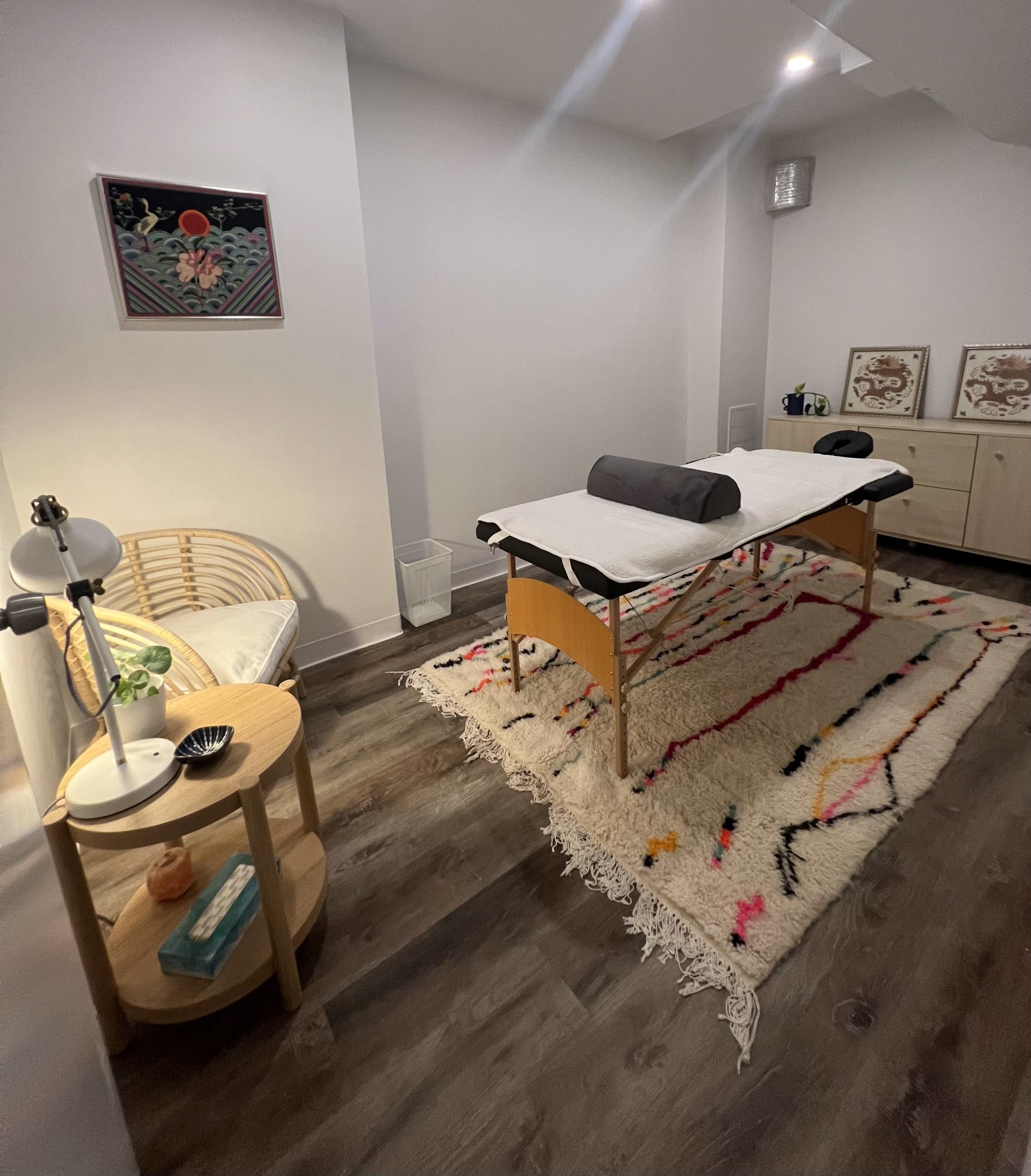 The image shows a serene massage therapy room featuring a treatment table, a comfortable chair, and decorative elements on the walls and floor.