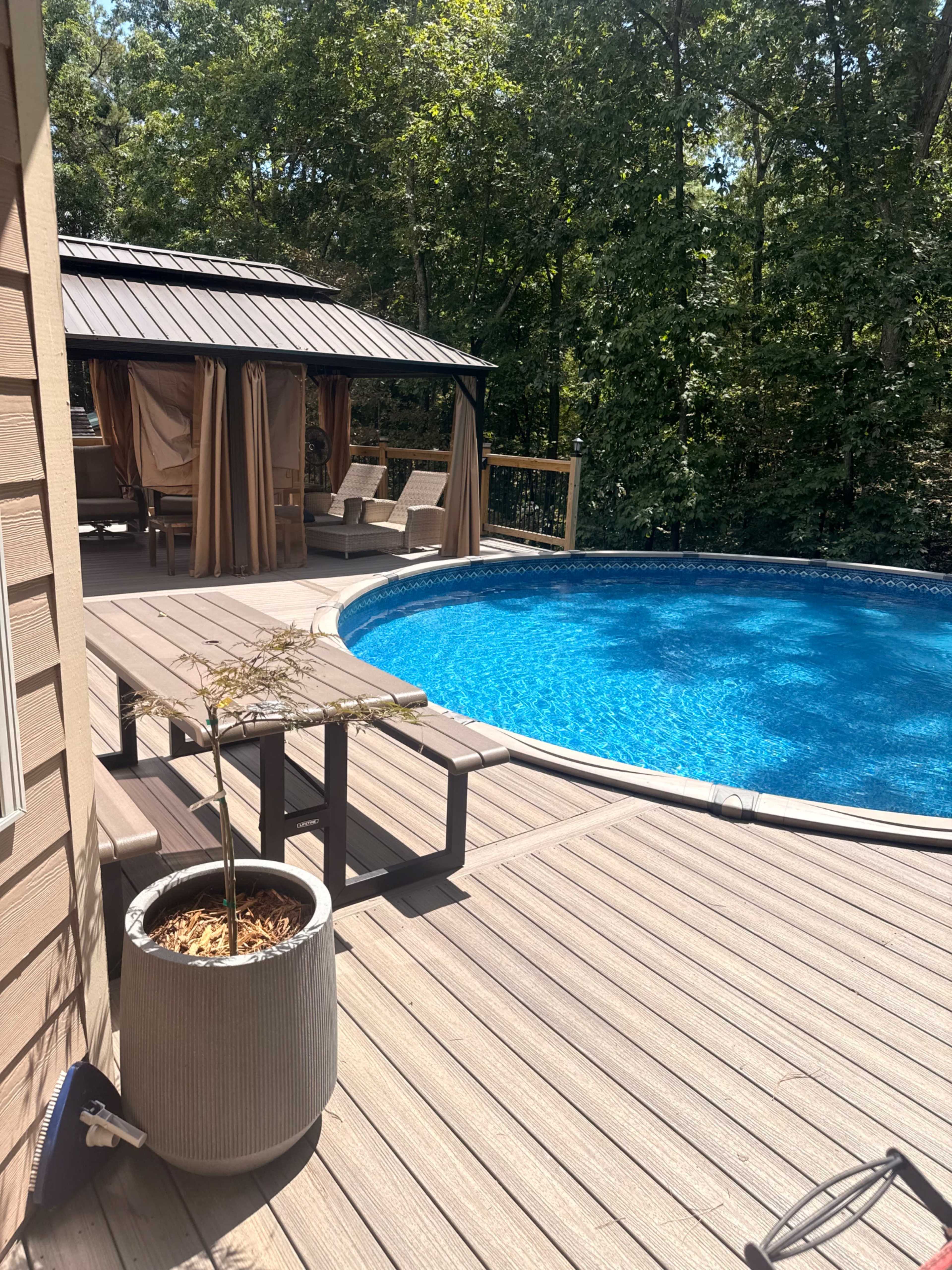 The image shows a poolside area featuring a circular pool, a wooden deck, outdoor seating under a gazebo, and lush trees in the background.