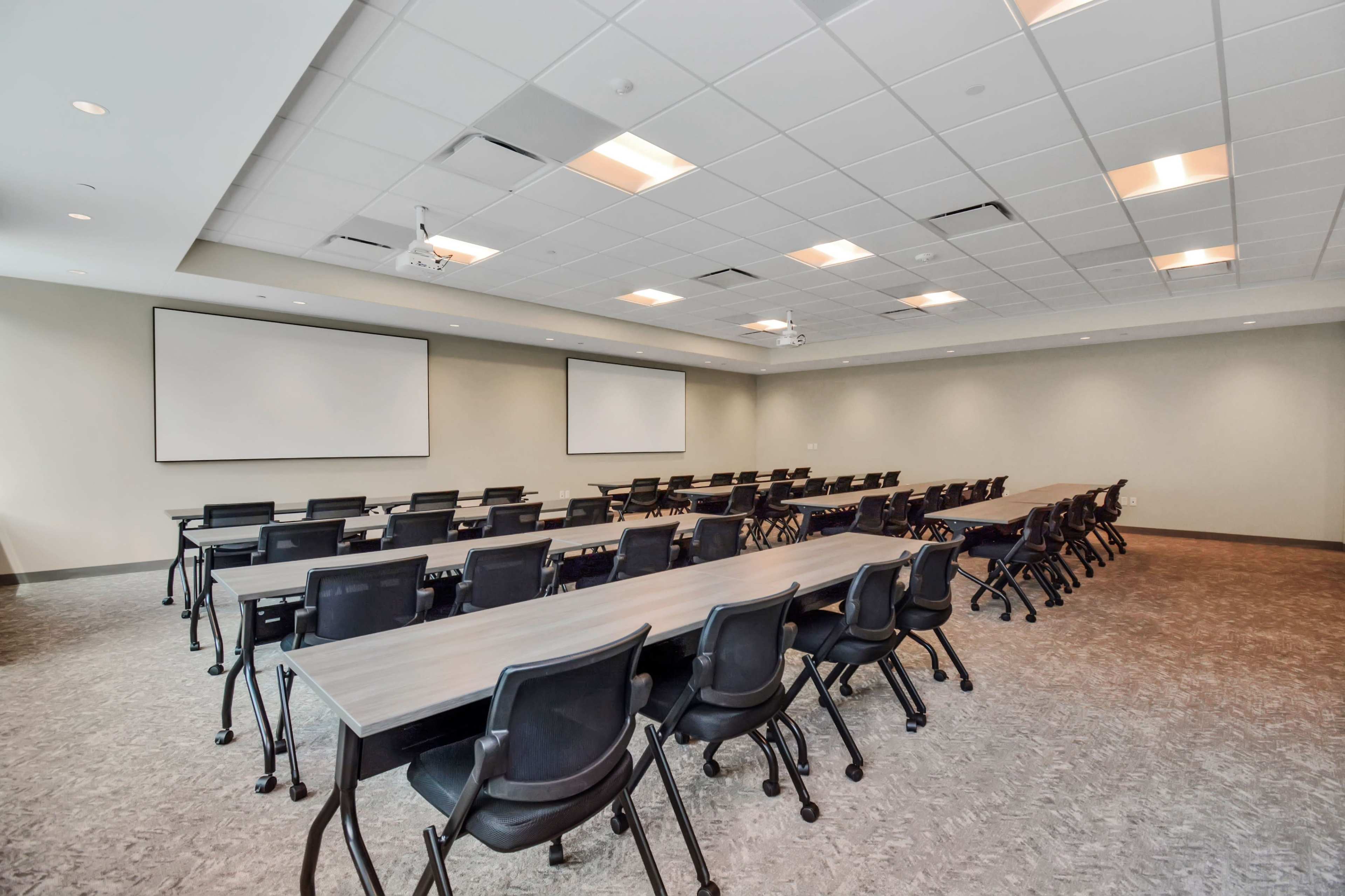 A large conference room features multiple rows of tables and chairs, with two blank projection screens on the front wall.
