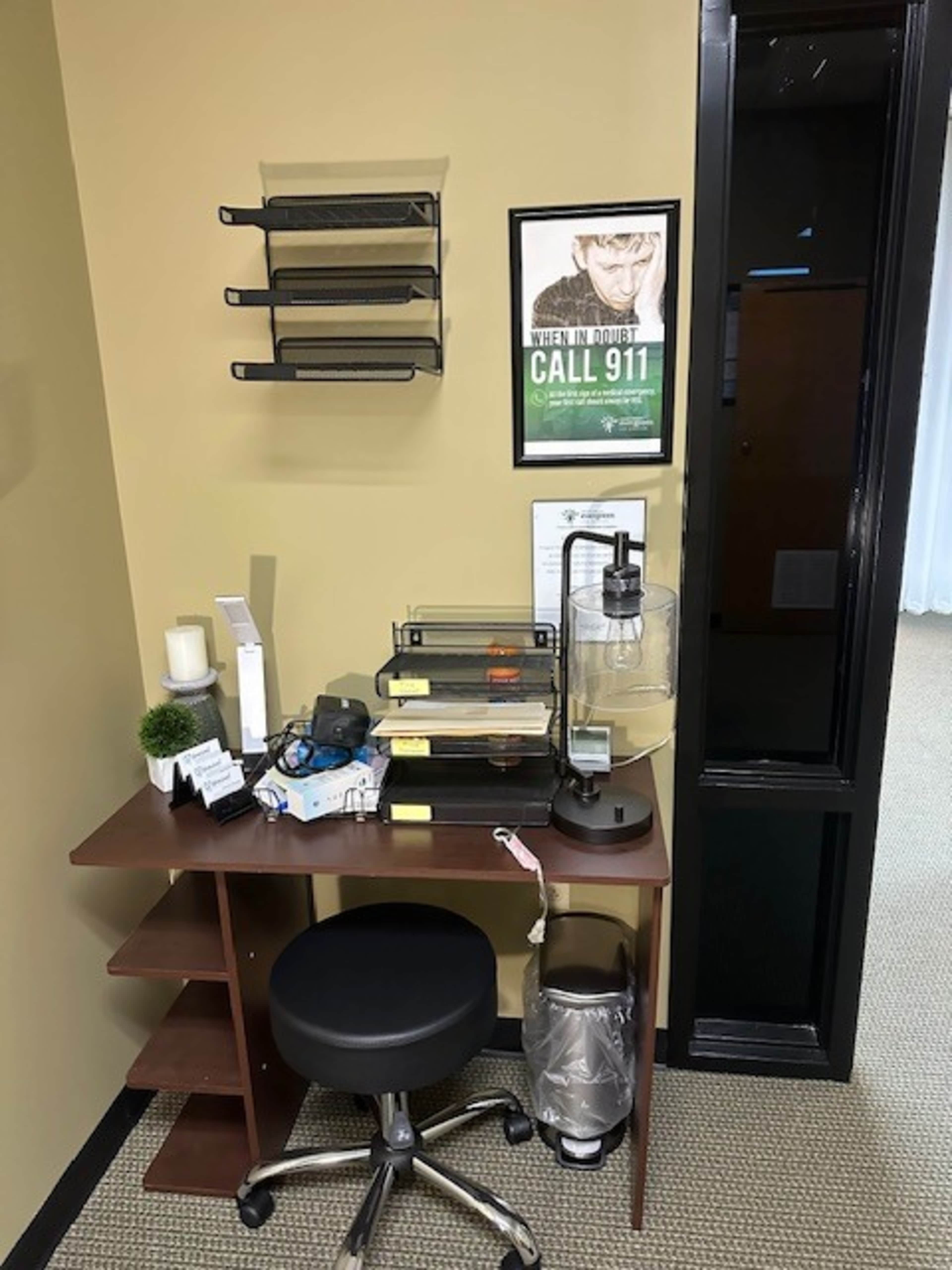 The image shows a small office corner with a desk, a rolling chair, a stack of shelves mounted on the wall, and a poster instructing to call 911, alongside various office supplies.
