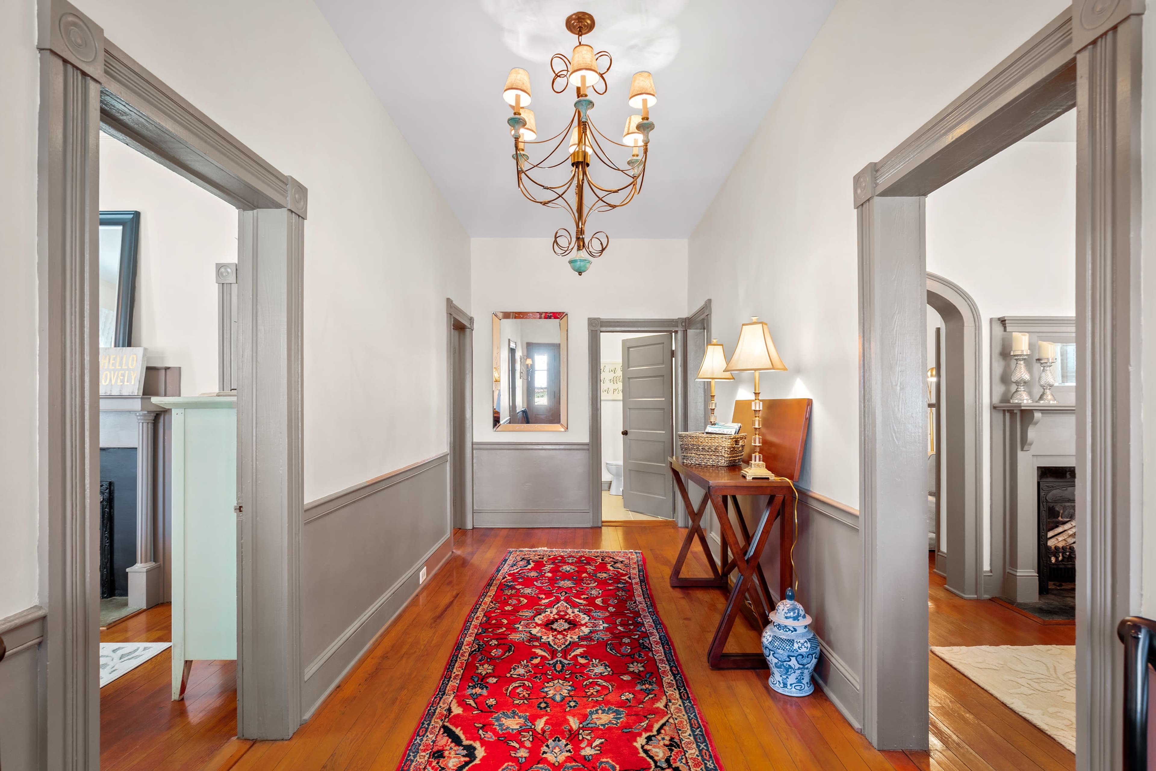 A well-lit hallway features a chandelier, a red runner rug, and doorways leading to adjacent rooms.