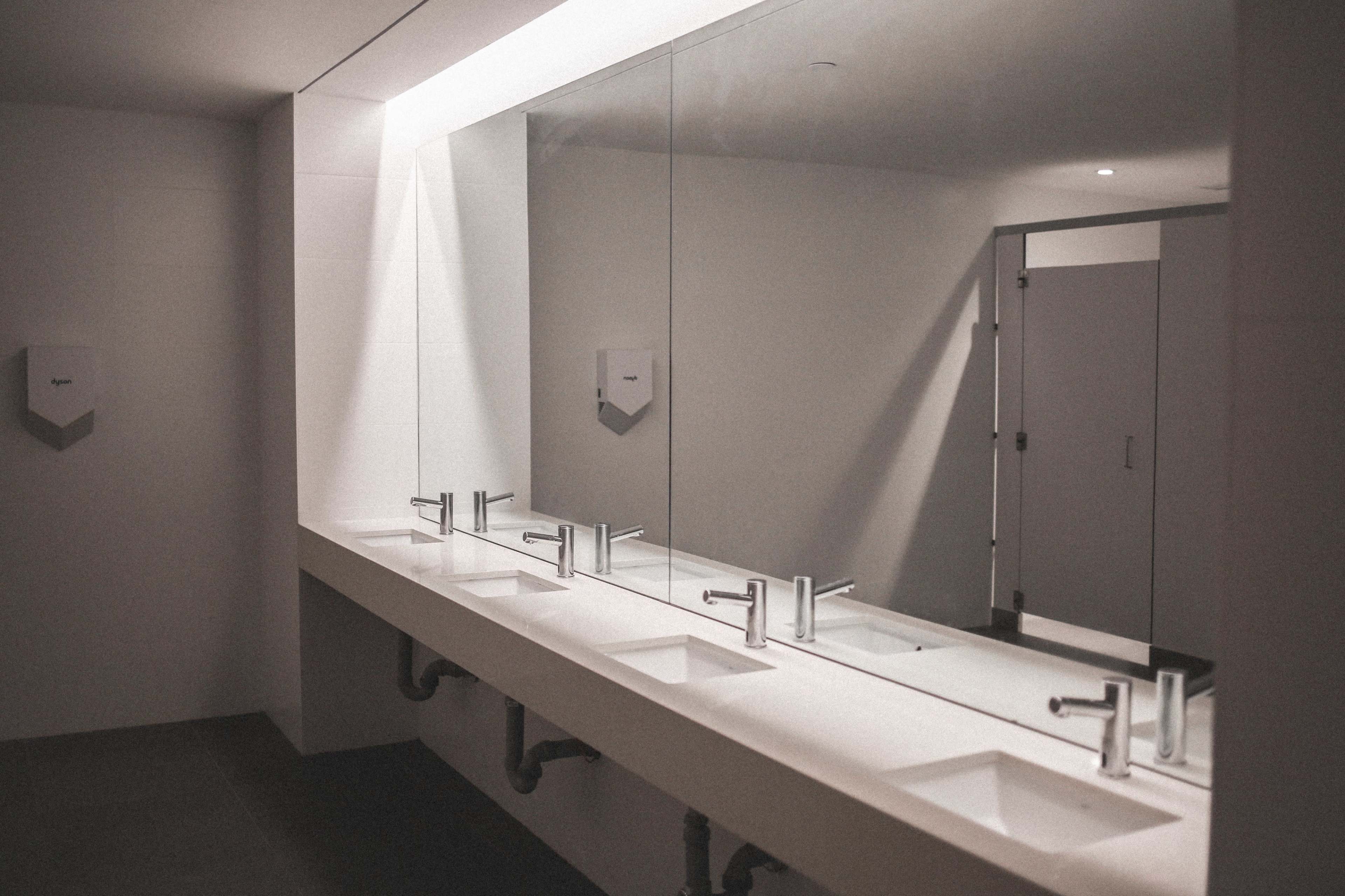 The image shows a modern restroom with multiple sinks and a large mirror reflecting the minimalist design.