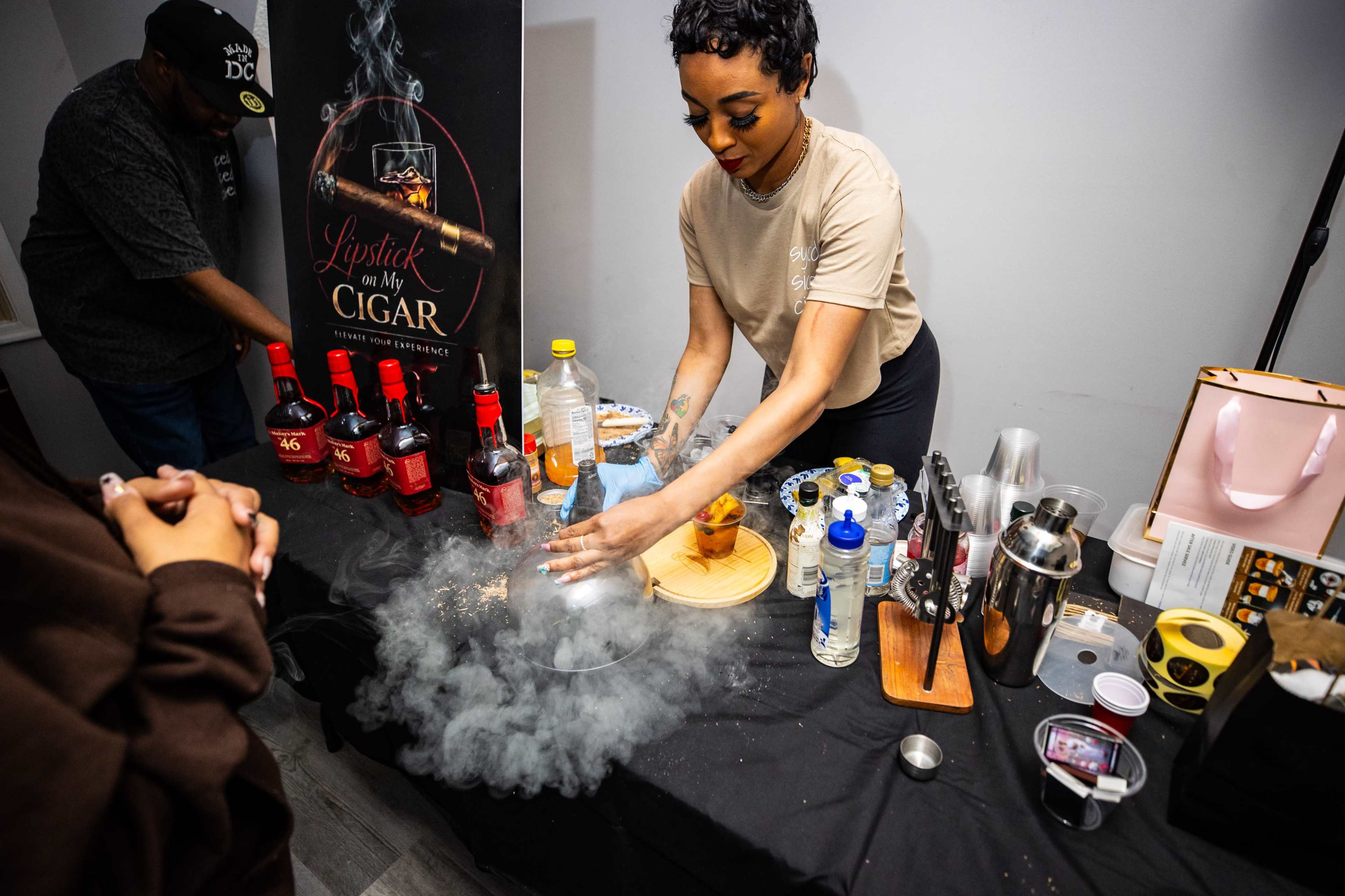 A woman prepares cocktails at a bar featuring various bottles and mixing tools, while vapor rises from a surface.
