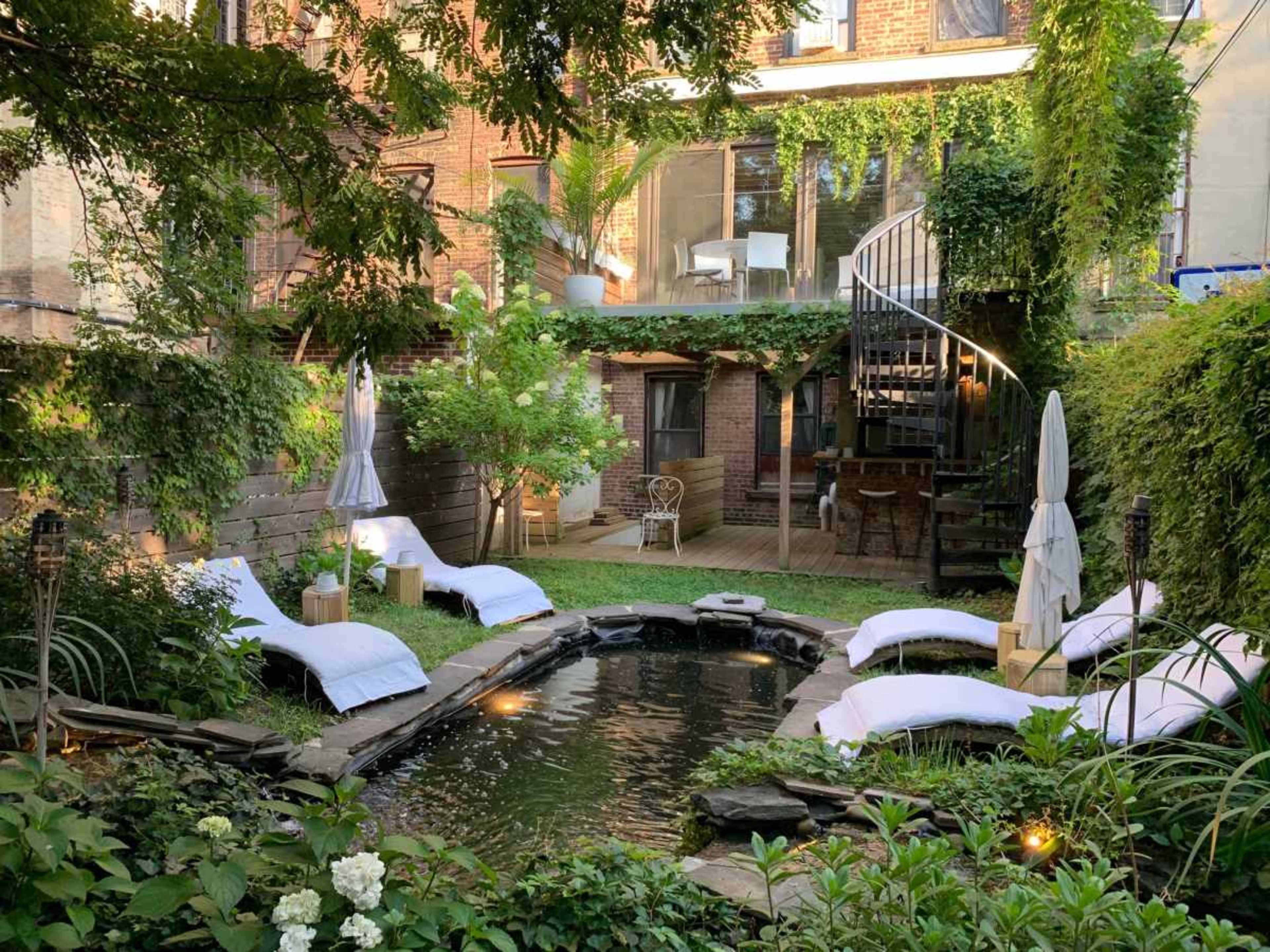 The image shows a backyard oasis with a small pond surrounded by lounge chairs, lush greenery, and a spiral staircase leading to a balcony.