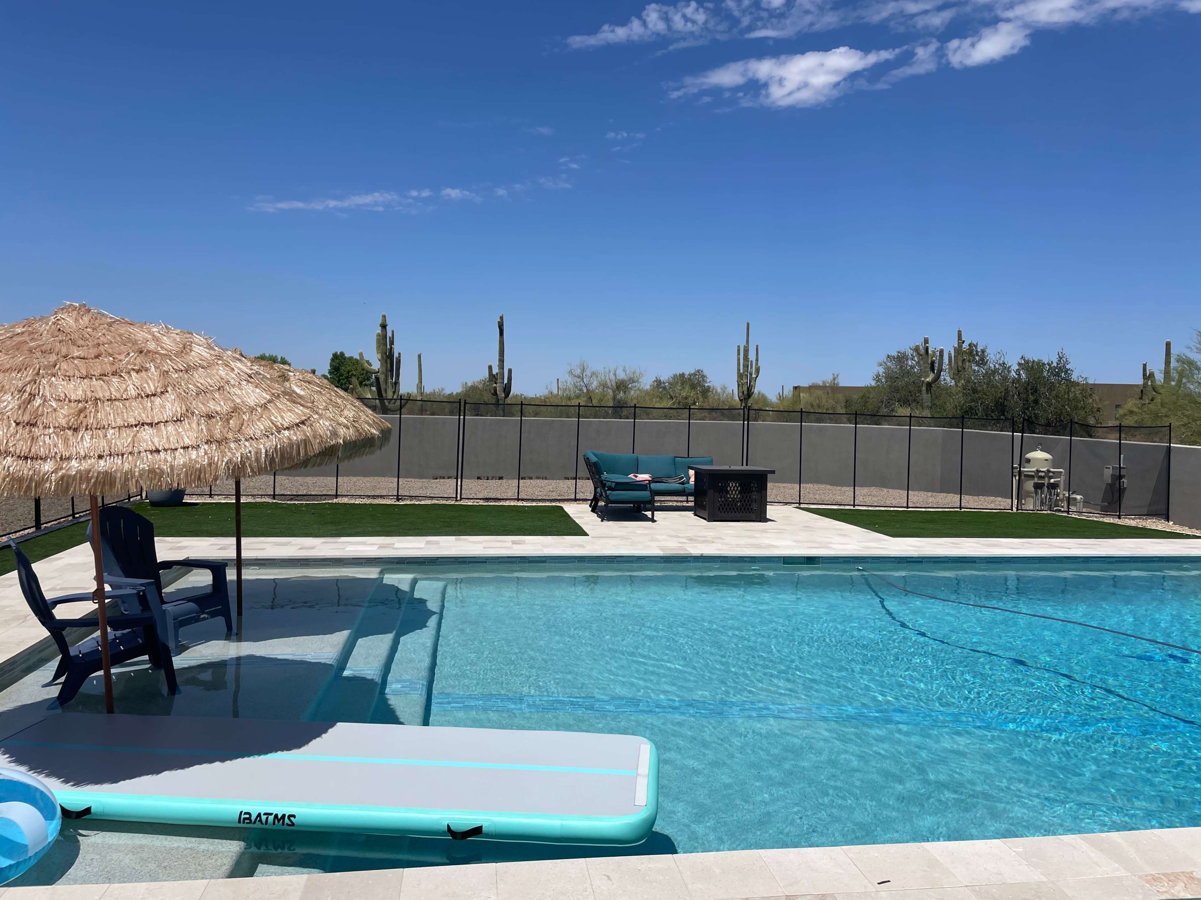 A swimming pool surrounded by a stone patio features a thatched umbrella, lounge chairs, and cacti in the background under a clear blue sky.