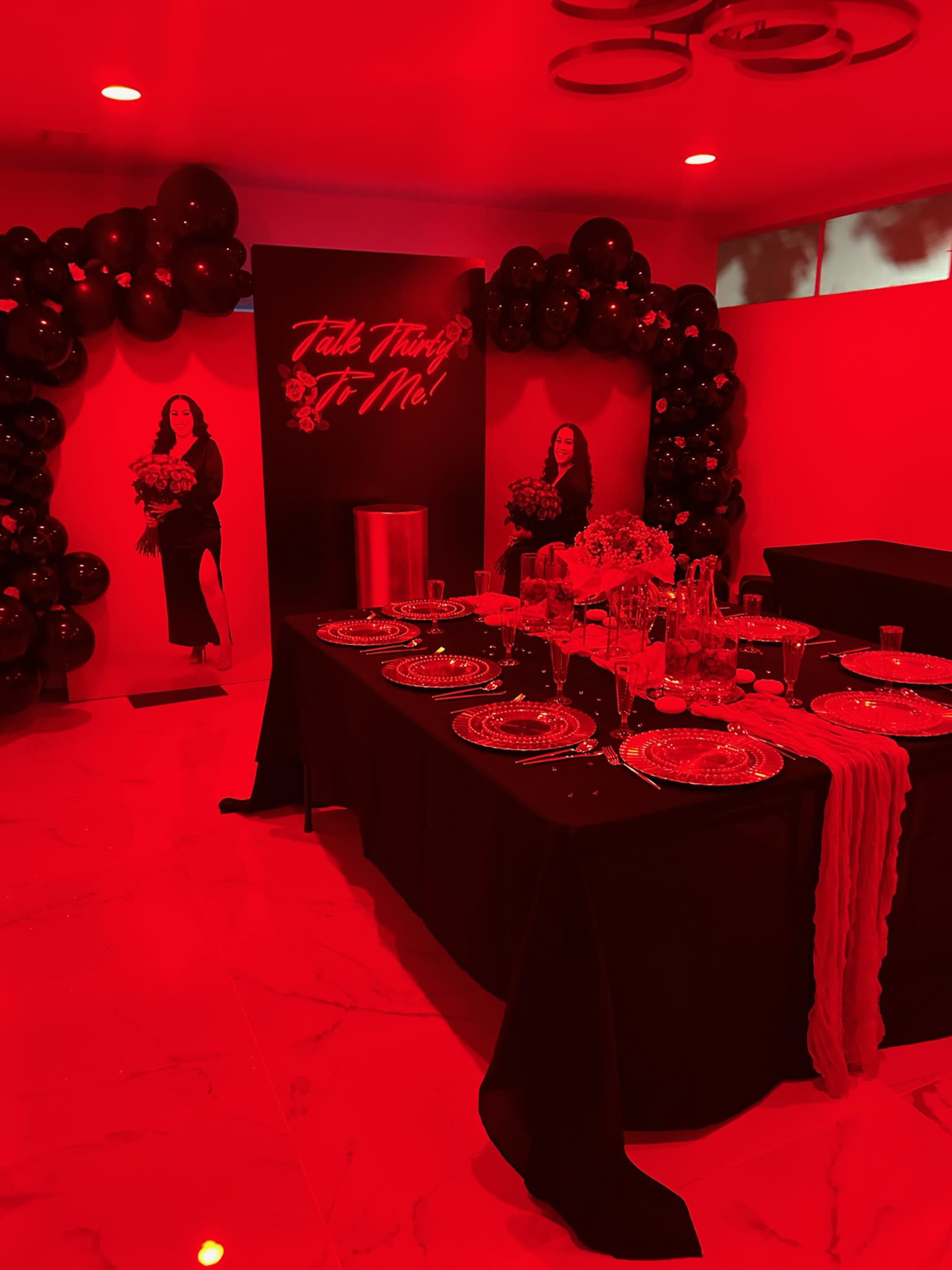 The image shows a decorated event space with a table set for dining, surrounded by black balloons and illuminated with red lighting.