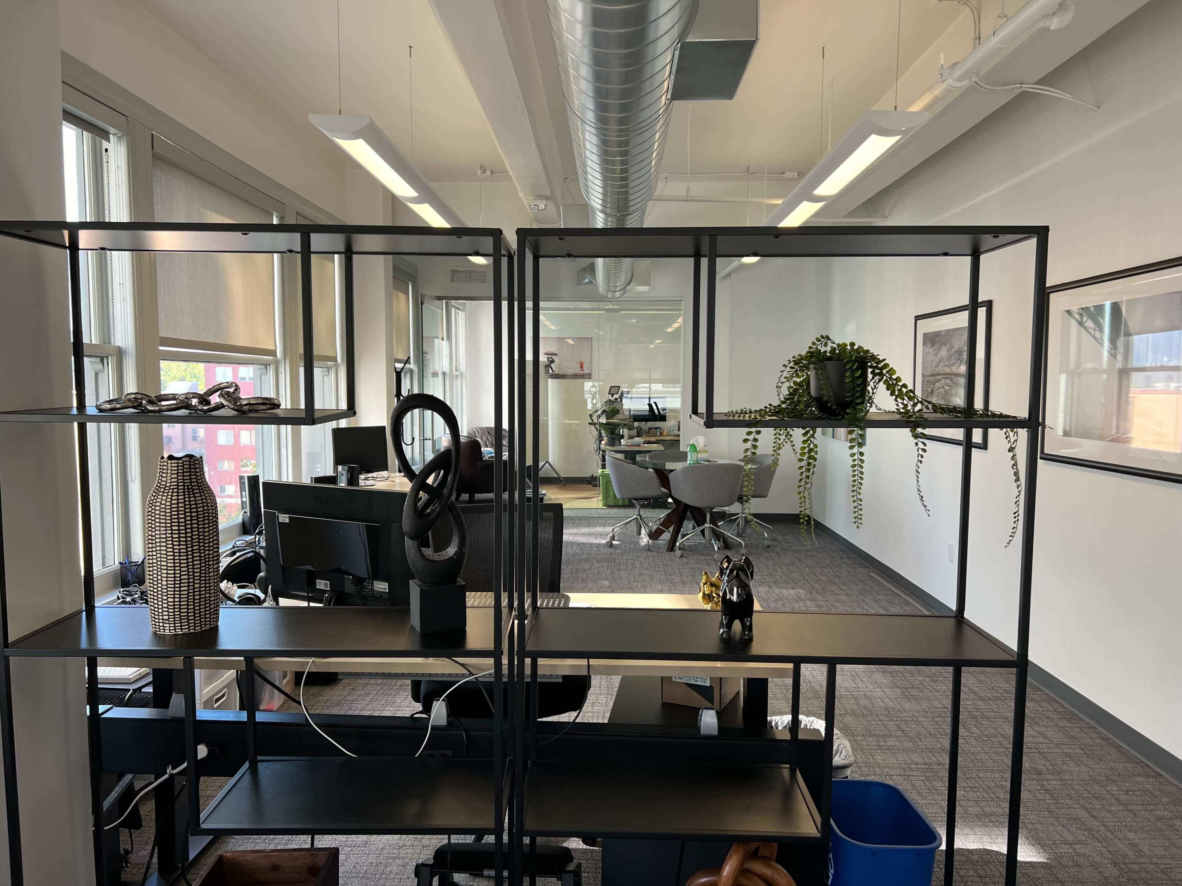 A modern office space with a black shelving unit in the foreground, displaying decorative items, and an office area with desks and chairs visible in the background.