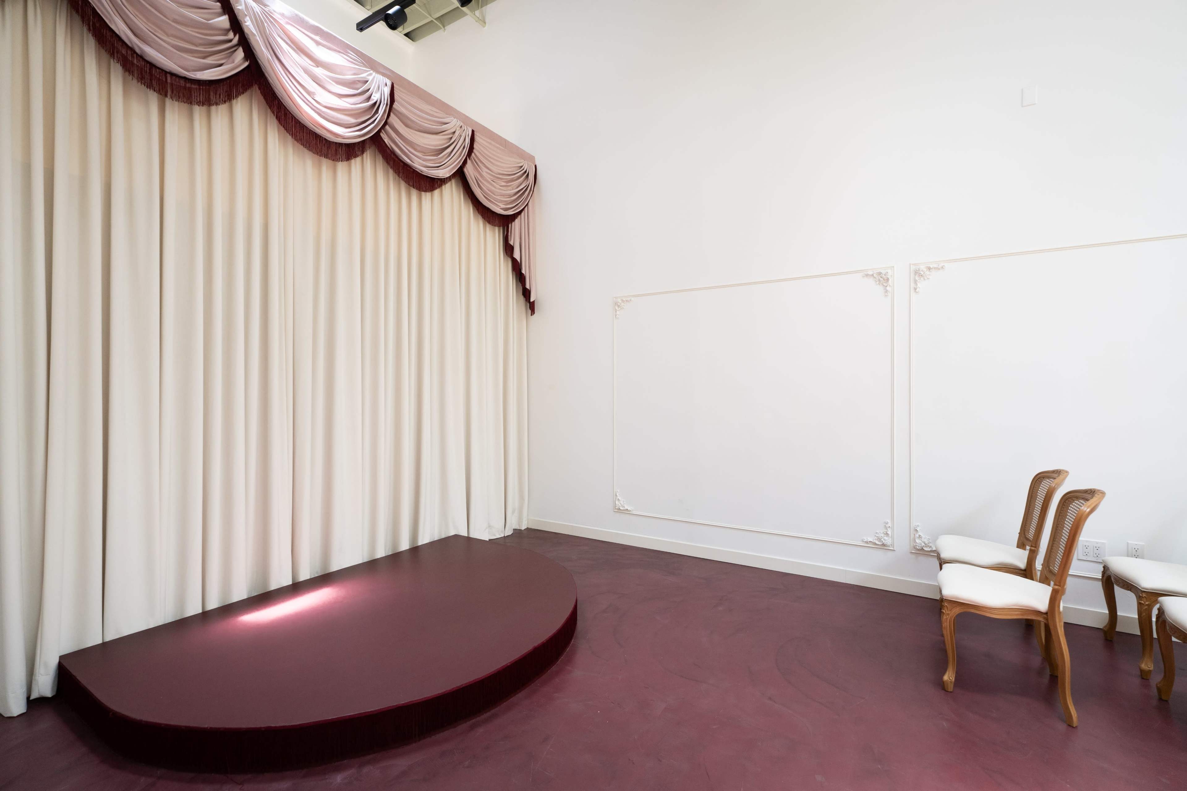 The image shows a stage with a curved maroon carpet, flanked by elegant drapes, and a blank wall next to two wooden chairs.
