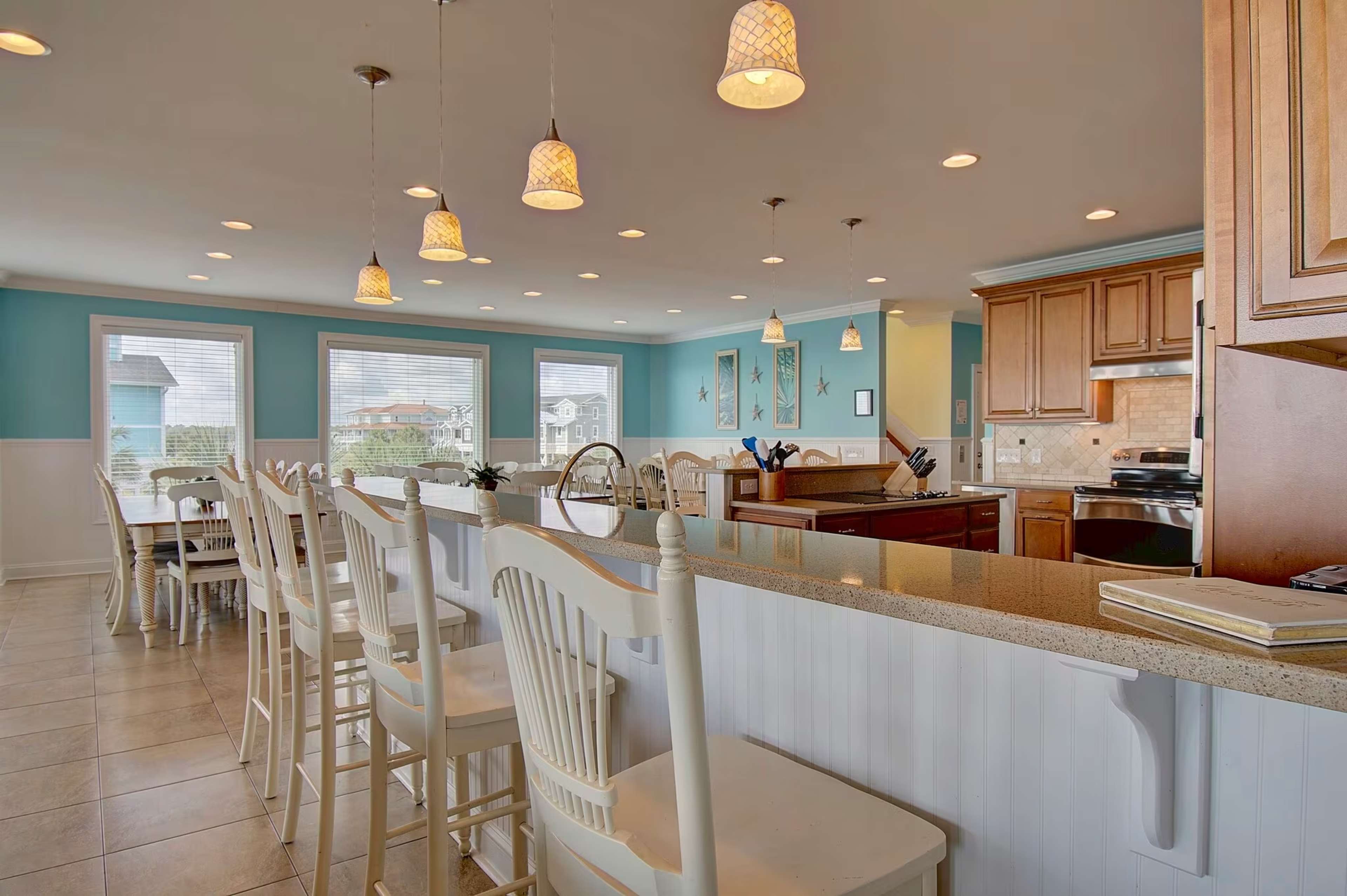 A spacious kitchen features a long bar with white stools, pendant lights, and a view of an adjacent dining area through large windows.