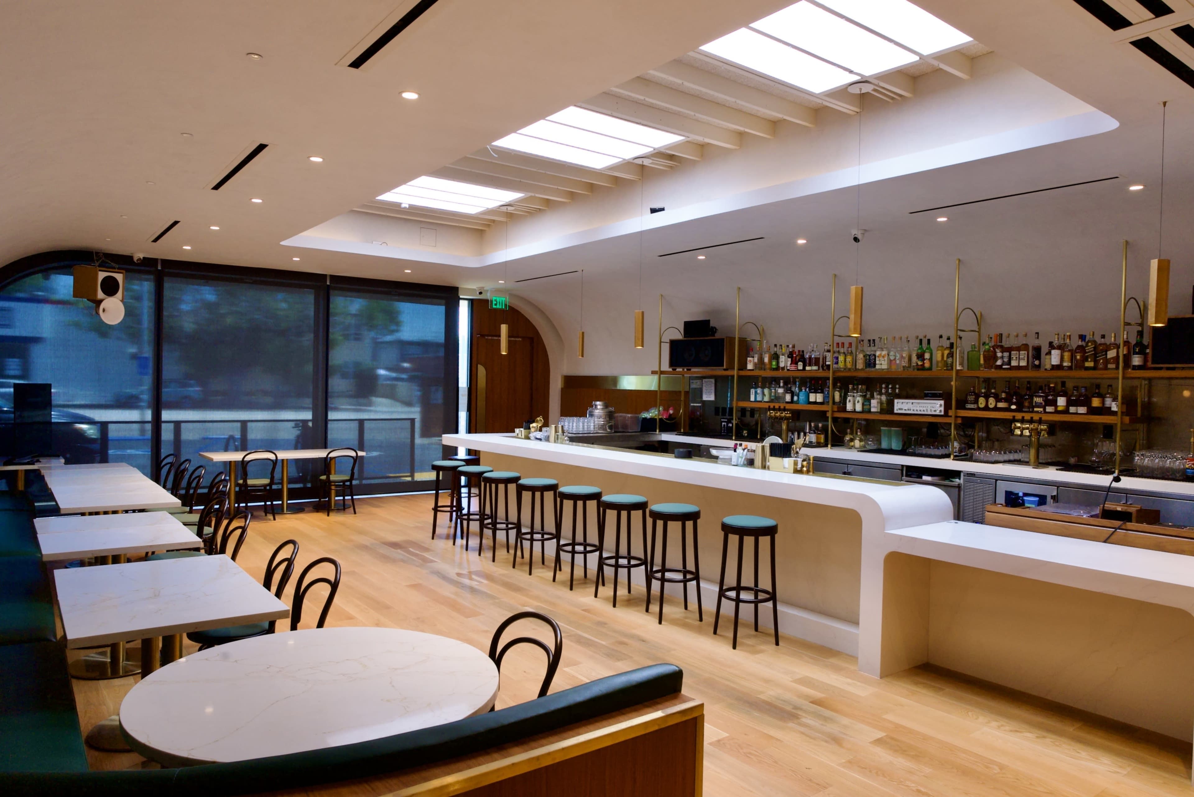 The image shows a modern bar area with a sleek white countertop, wooden floors, and large windows that provide natural light.
