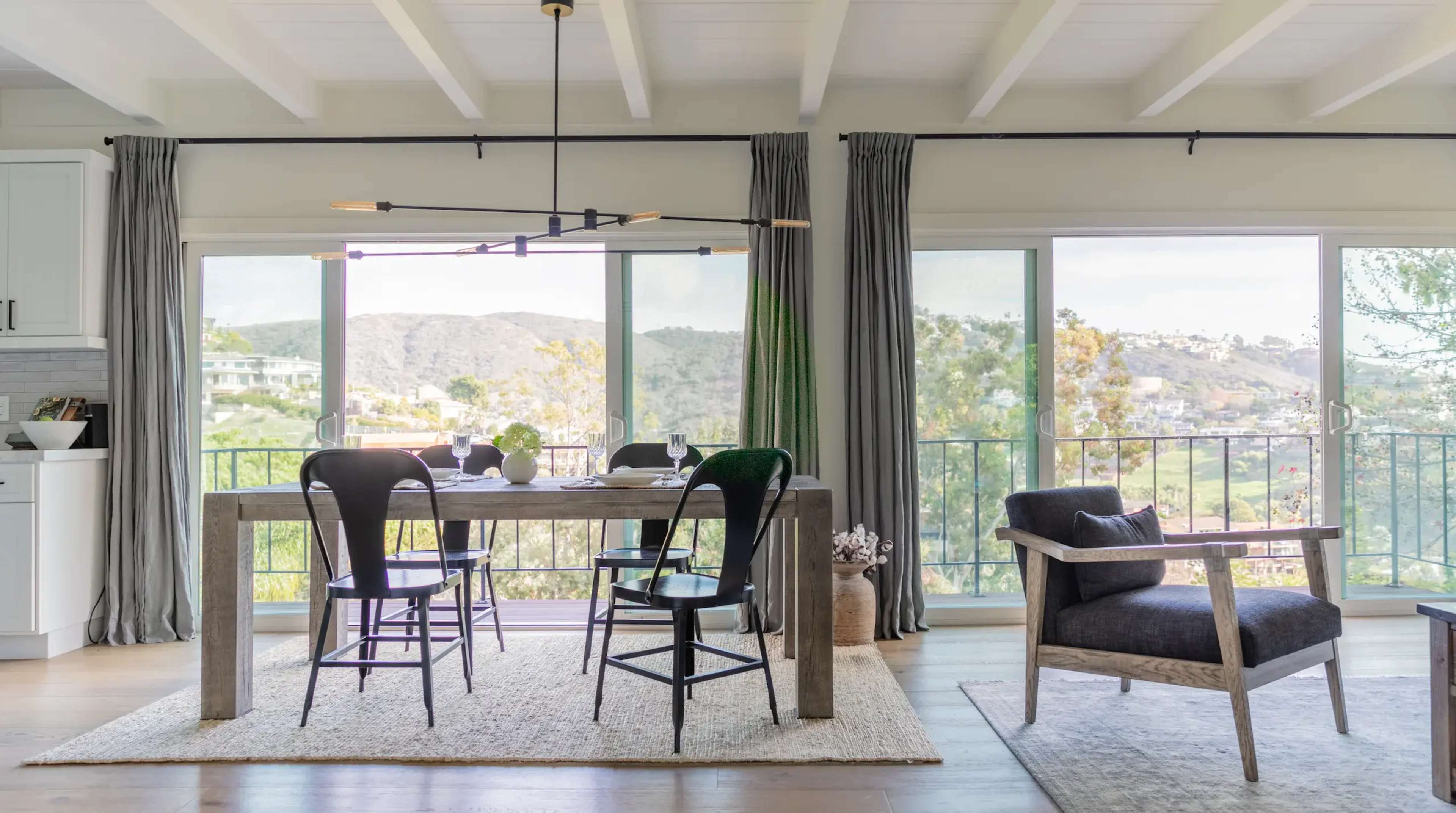 A modern dining area features a wooden table with two black chairs, surrounded by large windows that showcase a view of hills and greenery.