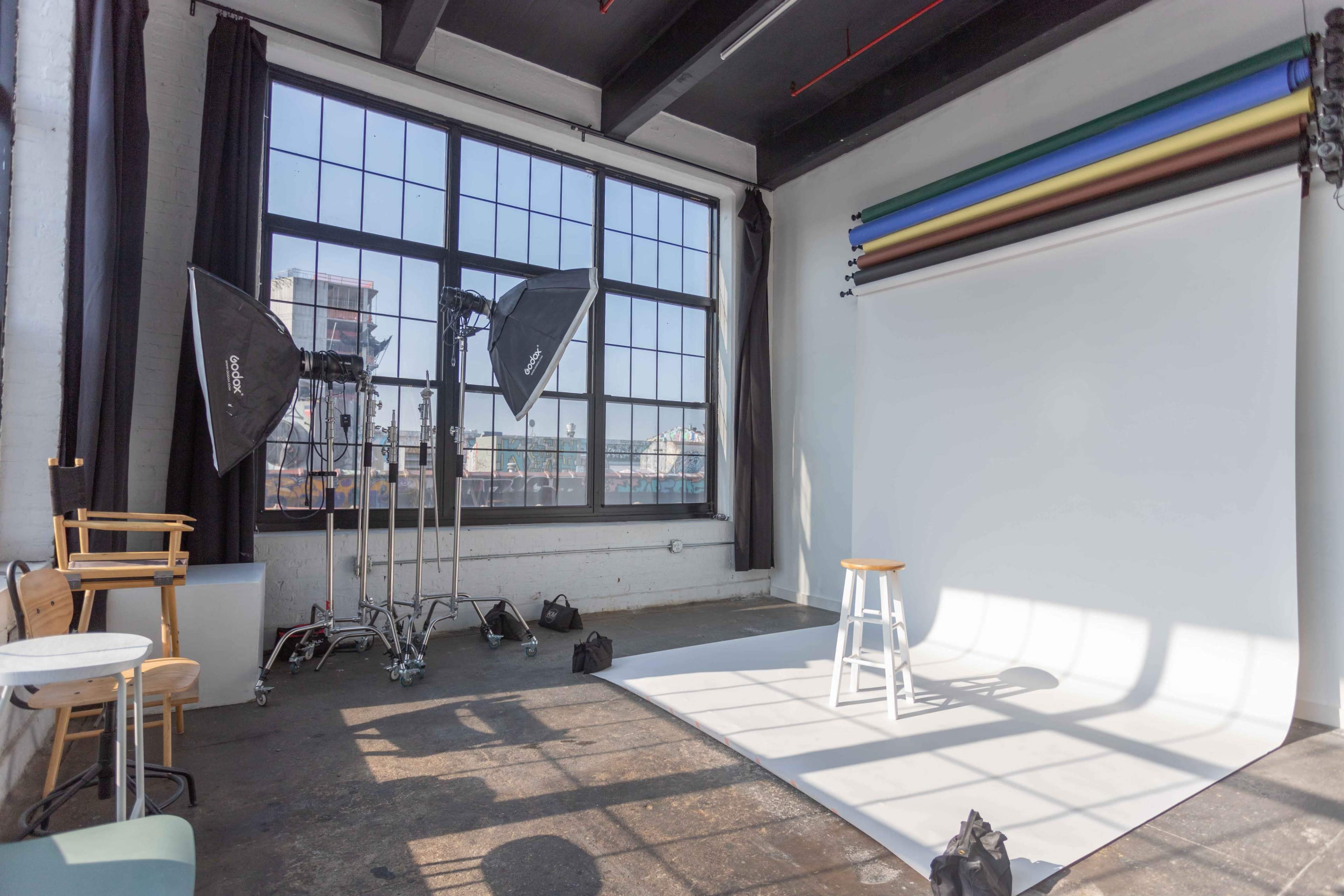 A photography studio featuring a white backdrop, a wooden stool, and several lighting equipment setups near large windows.