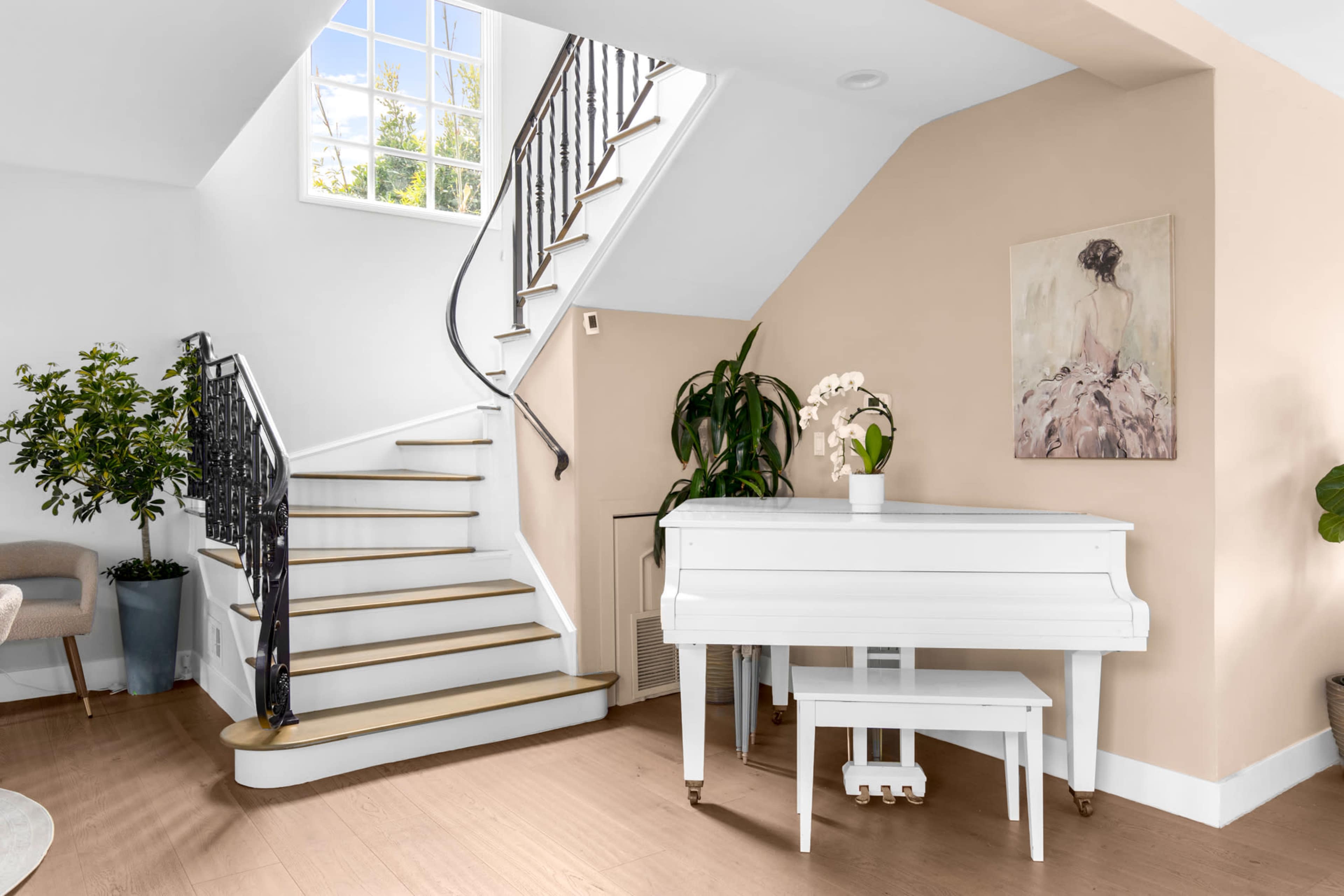 The image shows a bright foyer with a staircase, a white piano, and a painting on the wall, complemented by a potted plant in the corner.