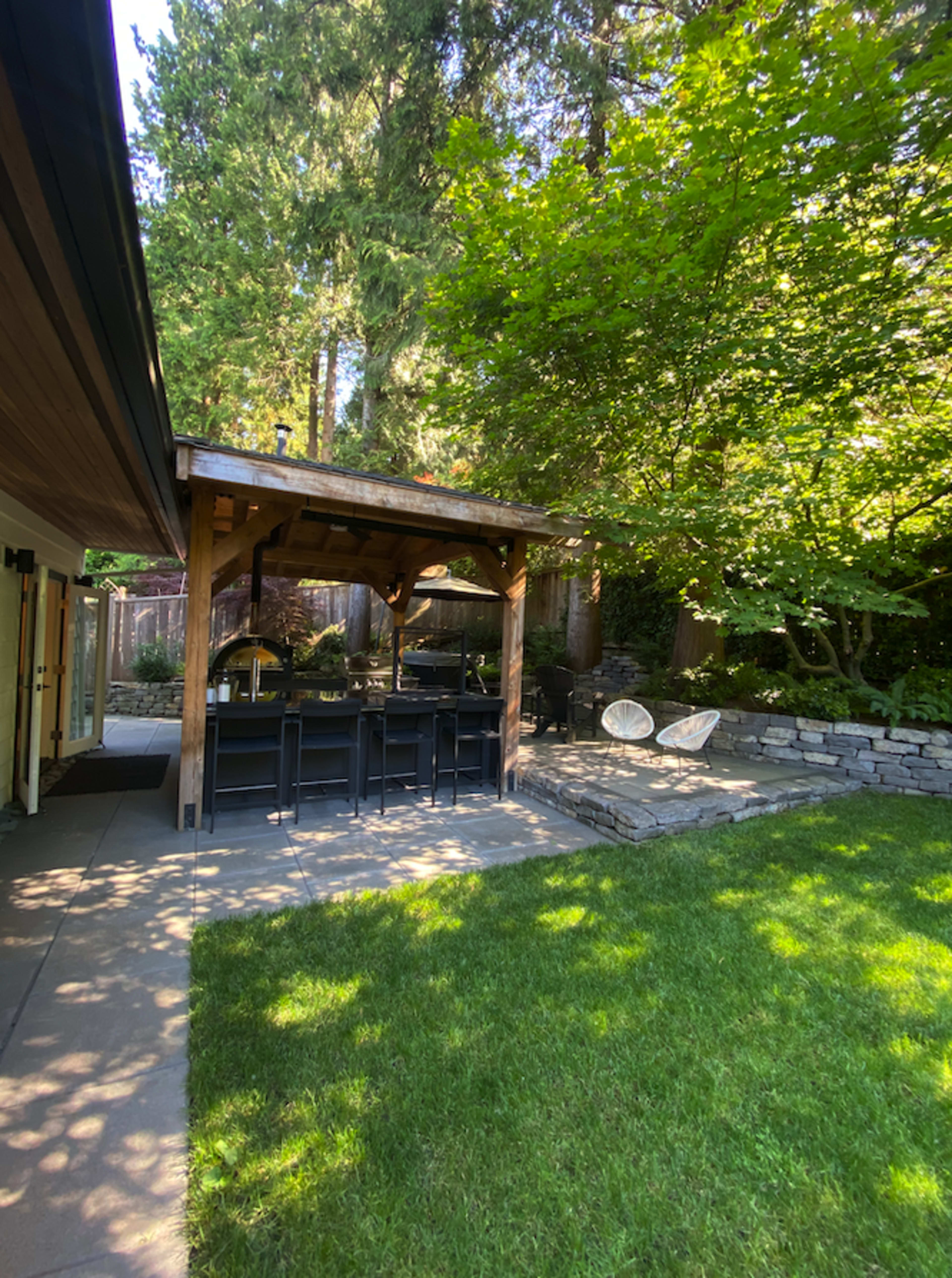 A wooden gazebo with seating is positioned next to a grassy area surrounded by trees and stone landscaping.