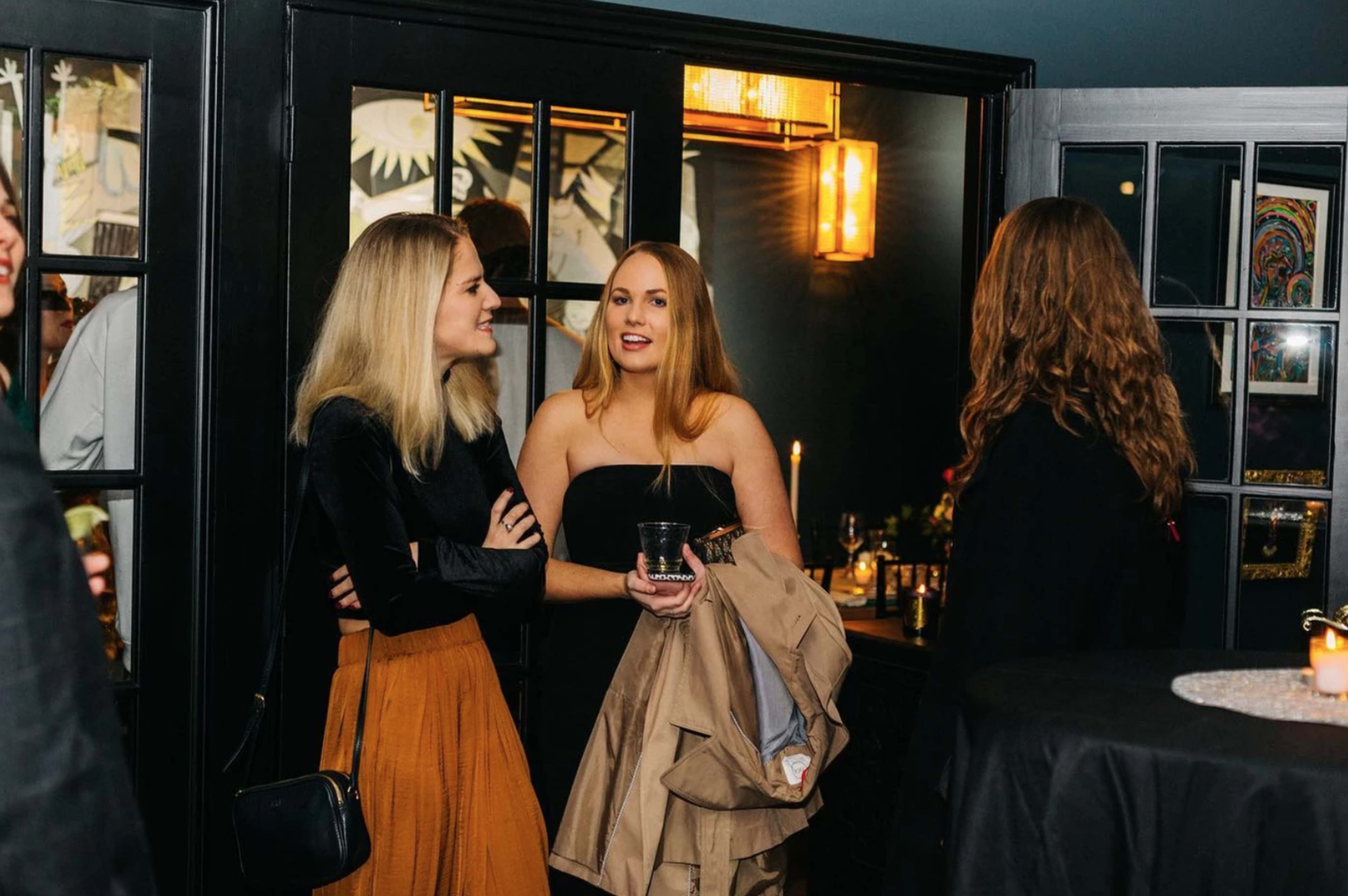 Two women are talking in a dimly lit room, one holding a drink and a coat while the other listens.