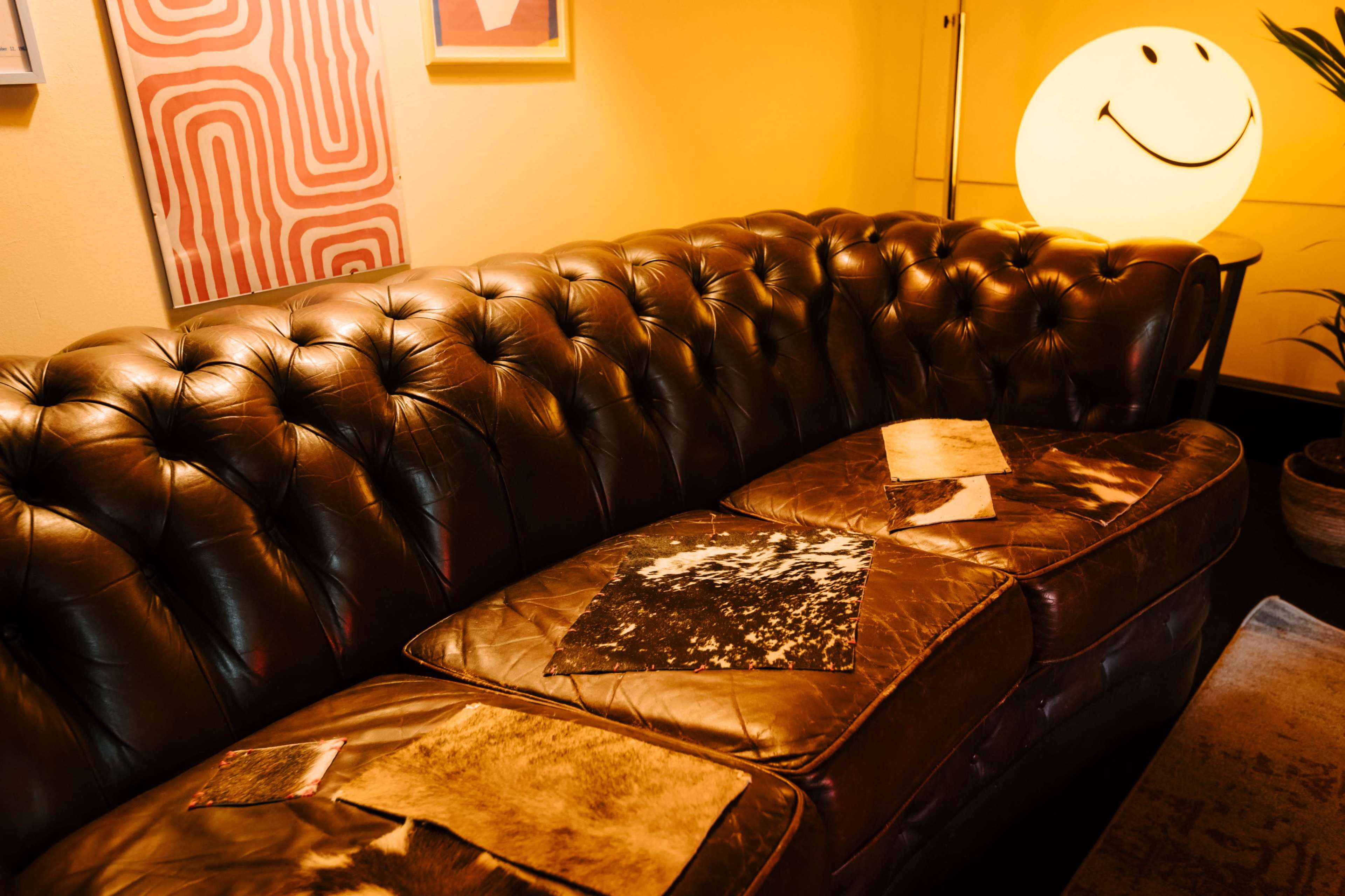 A brown leather tufted sofa is positioned in a warm-lit room, adorned with abstract art and featuring a round light fixture shaped like a smiling face.