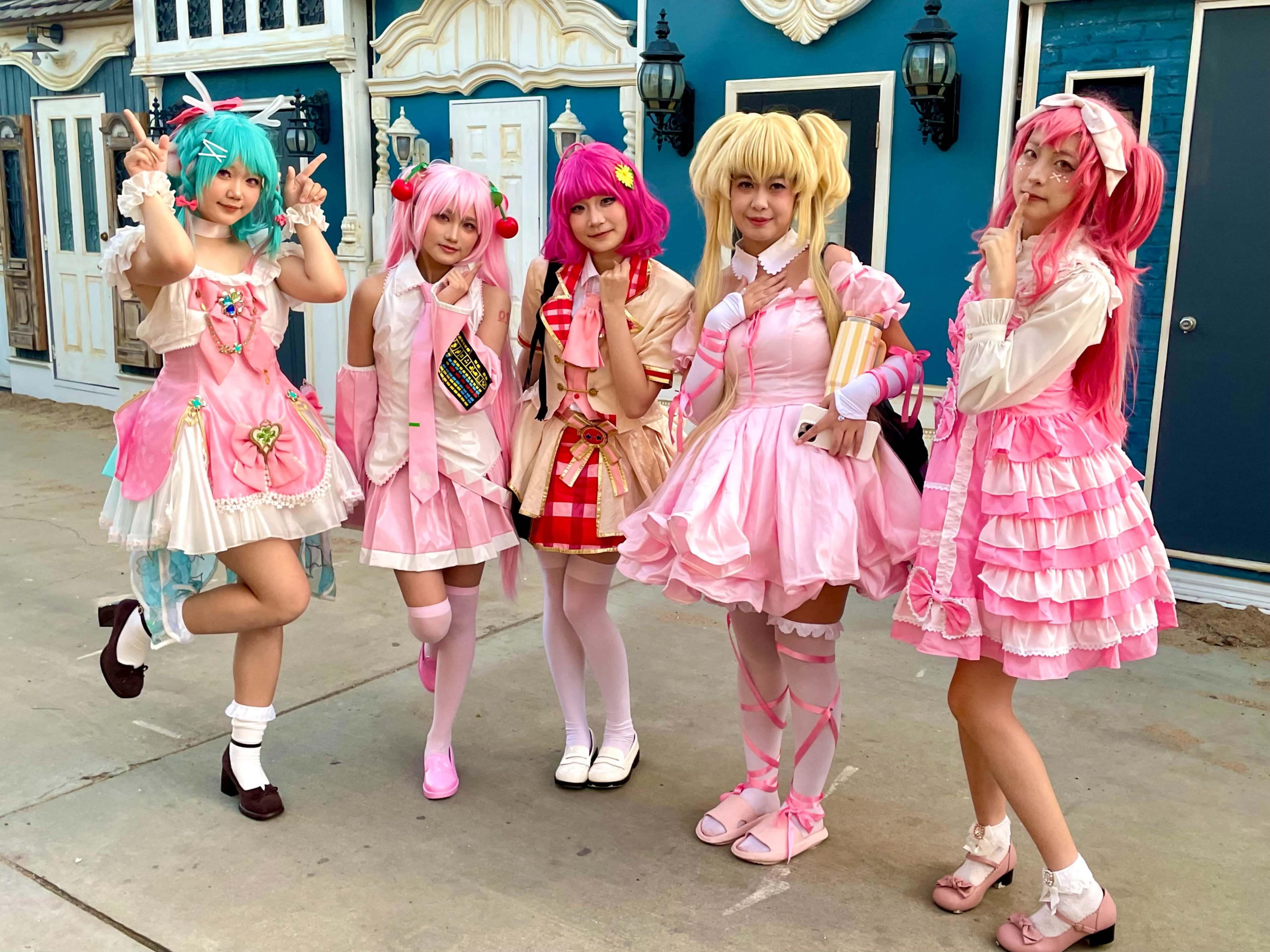 Five individuals dressed in colorful, coordinated outfits pose together in a playful manner against a backdrop of pastel-colored buildings.