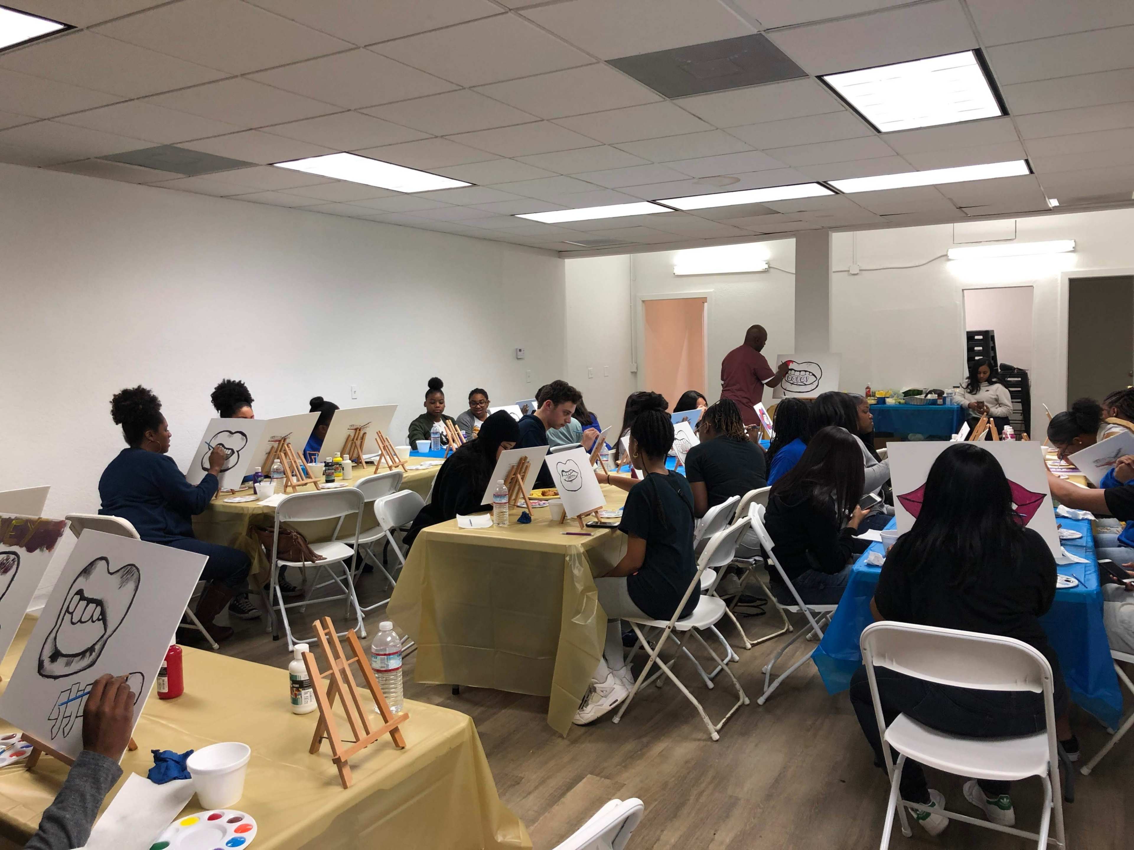 A group of people sit at tables in a well-lit room, participating in a painting class, with canvases displaying images in front of them.