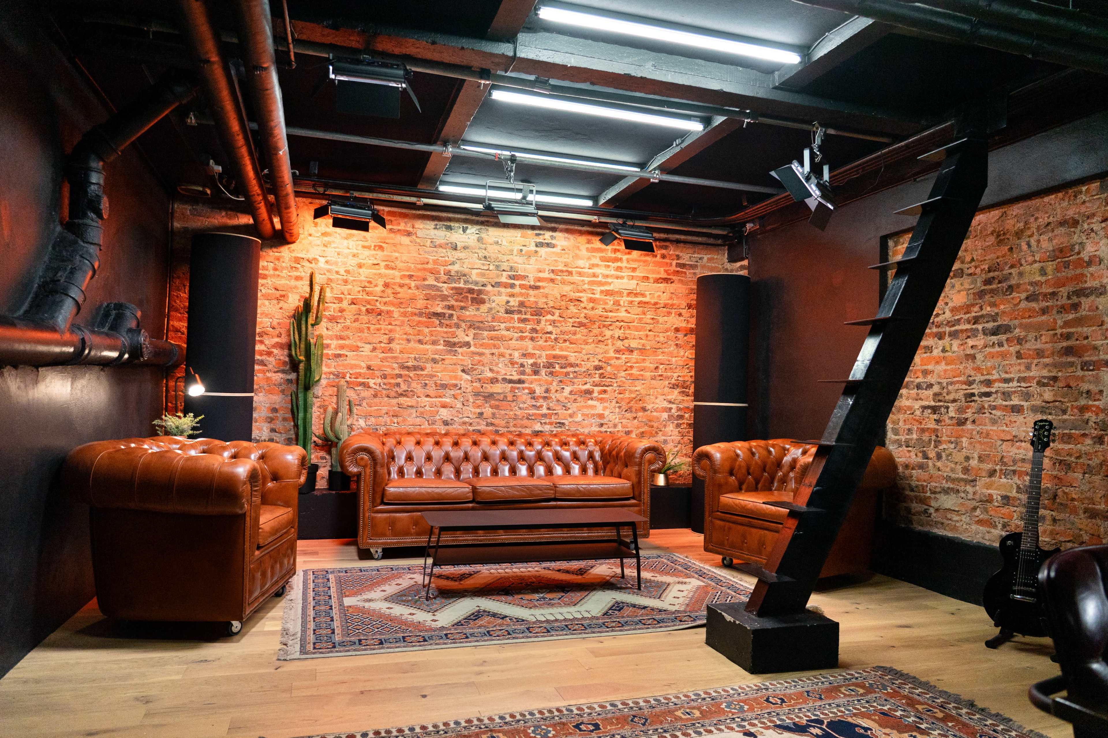 The image shows a cozy room with exposed brick walls, two brown leather sofas, a coffee table, and a guitar leaning against a column.