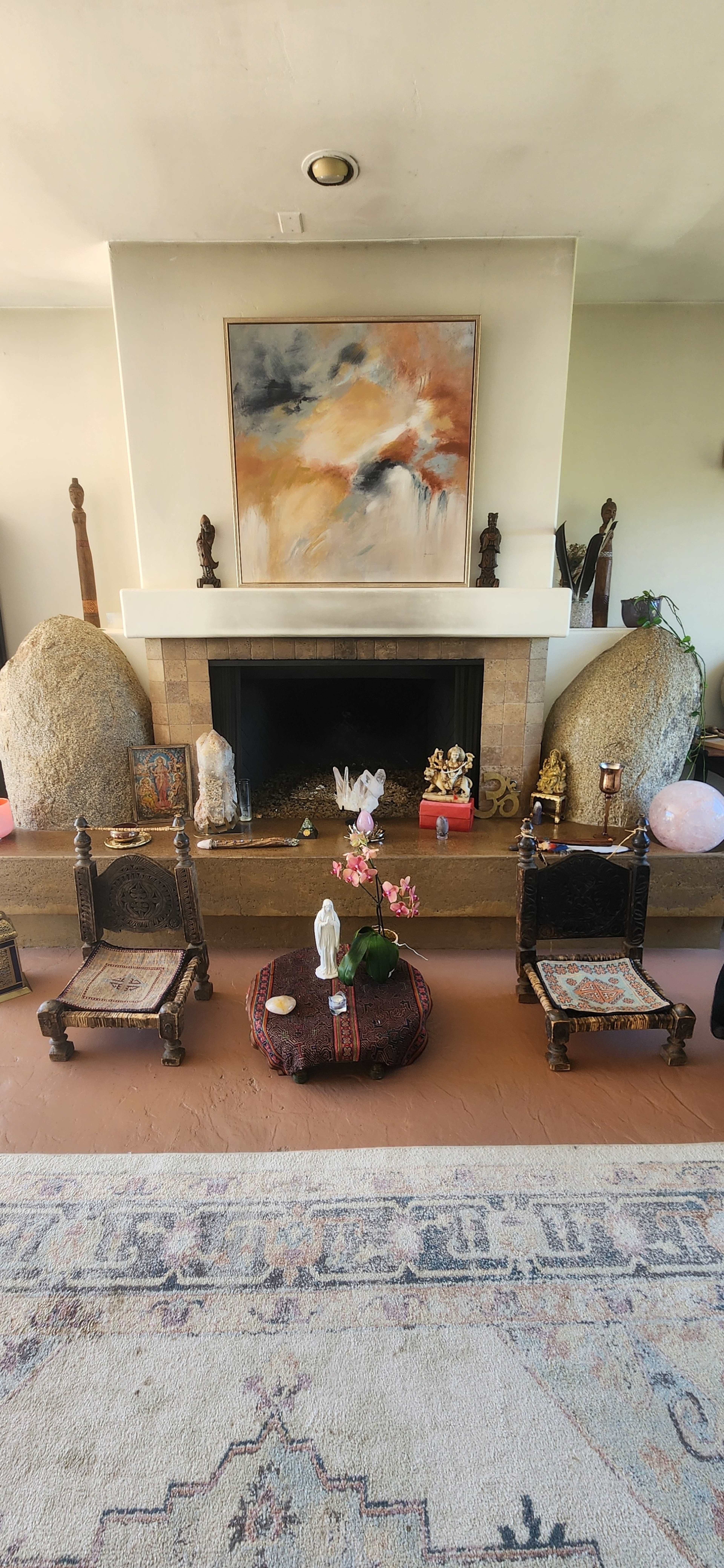 A living room with a large stone fireplace, two decorative stones on either side, wooden chairs, and a small table with a plant and various decorative items.