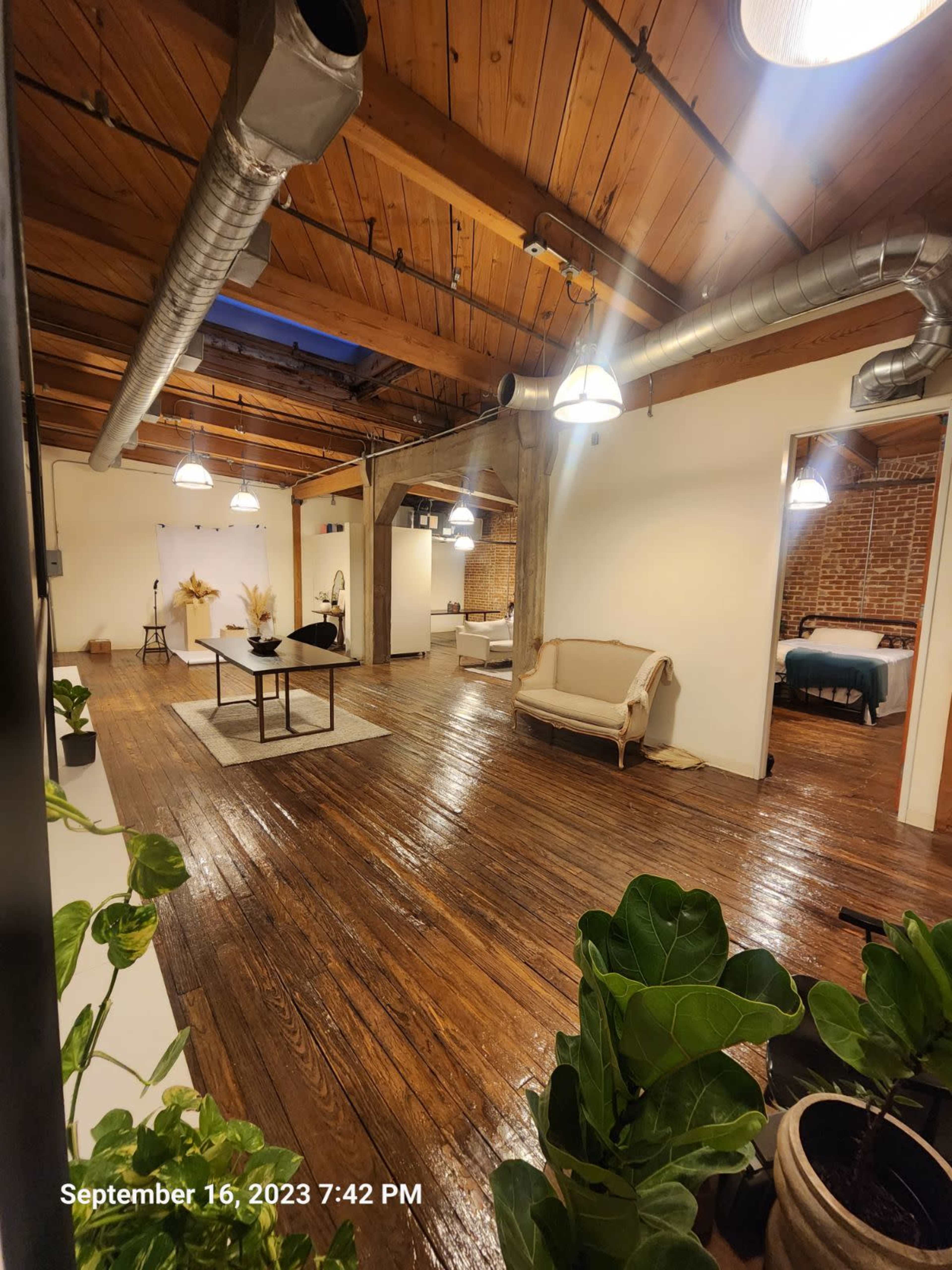 A spacious, industrial-style interior with wooden floors, exposed beams, and a mix of seating areas and decorative plants.