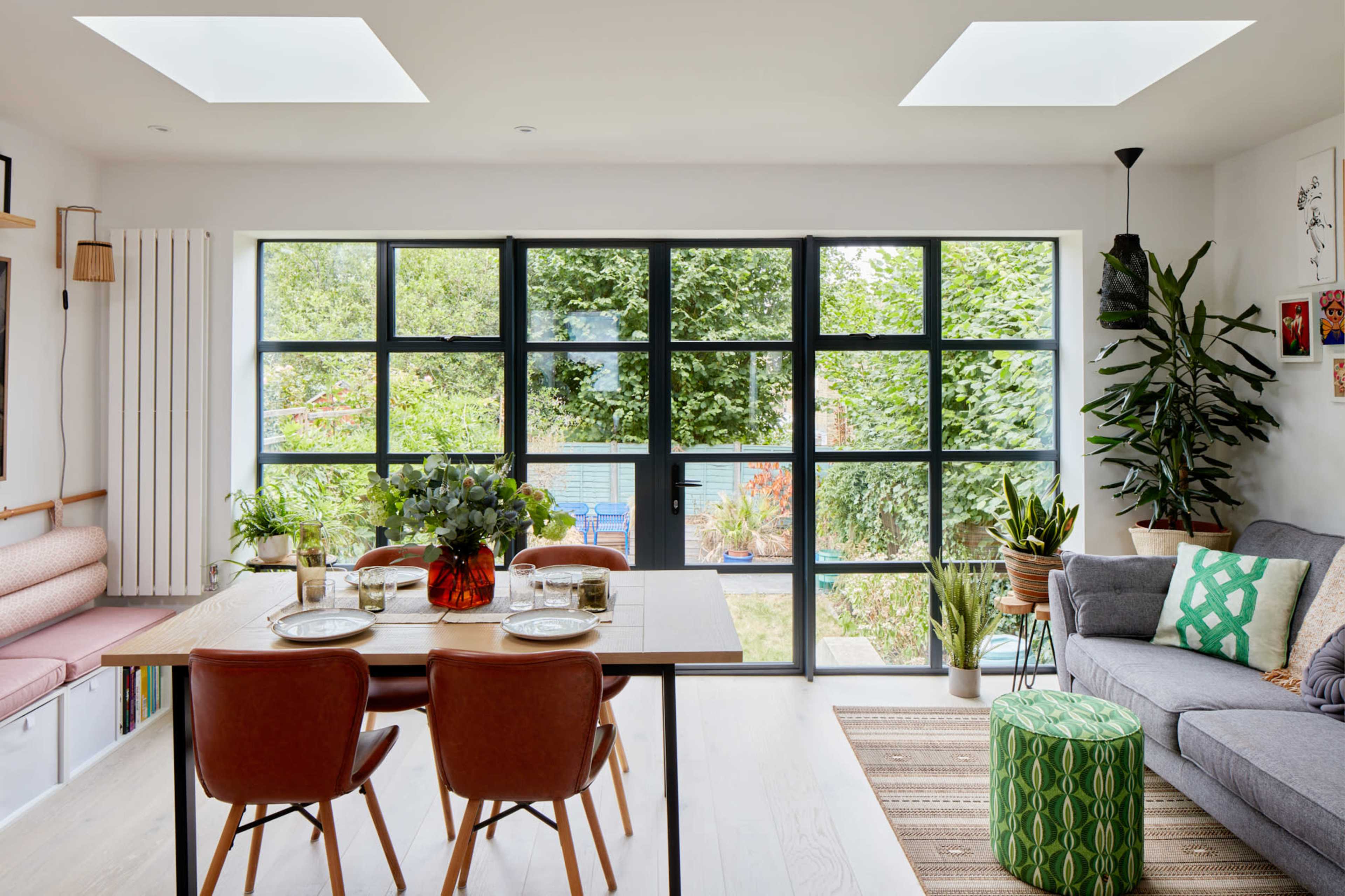 A modern dining area features a table set for four, surrounded by large windows that overlook a garden, with a sofa and plants in the adjacent living space.