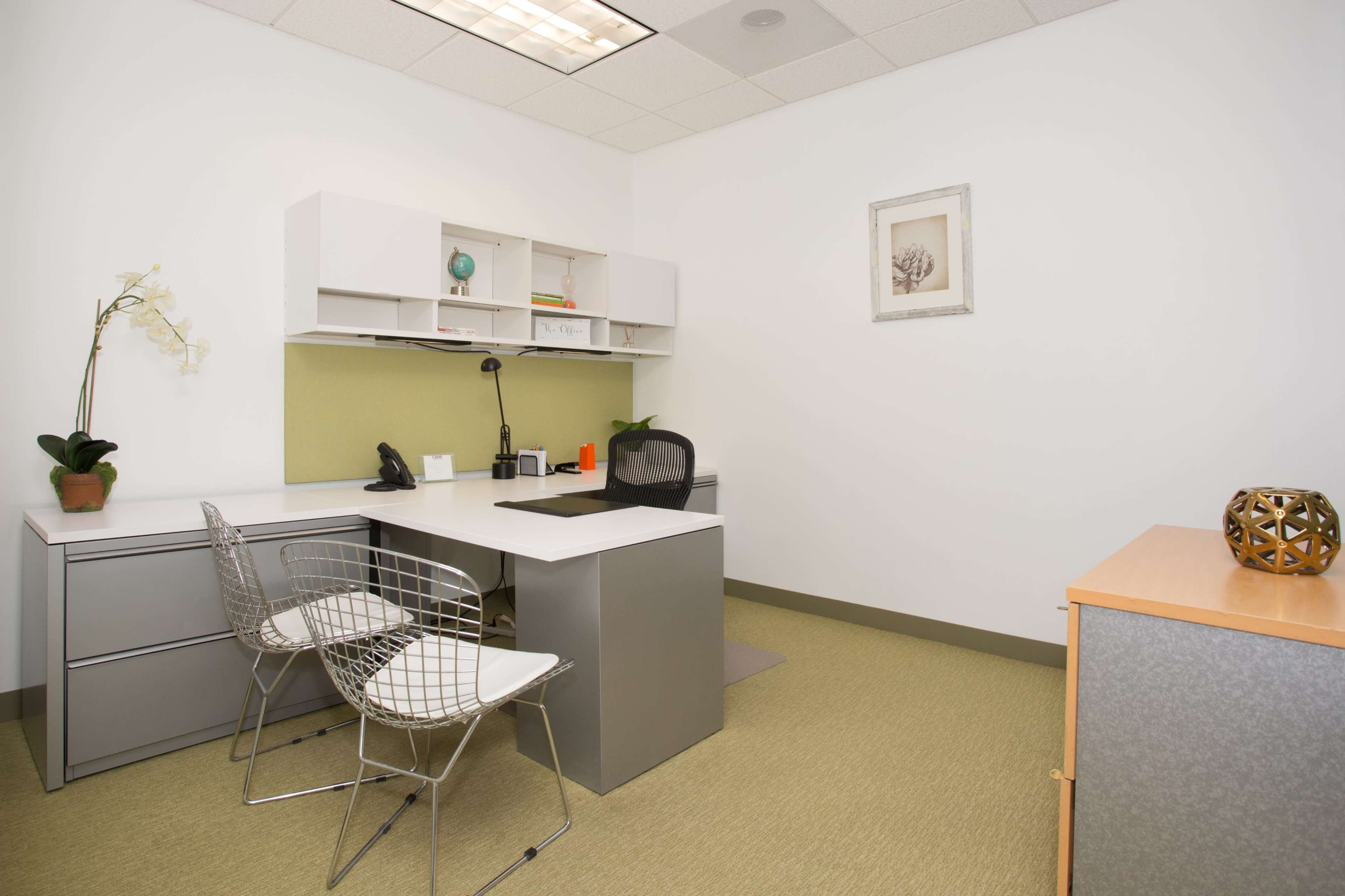 The image shows a modern office layout with a desk, two wire-frame chairs, and storage cabinets against a light-colored wall.