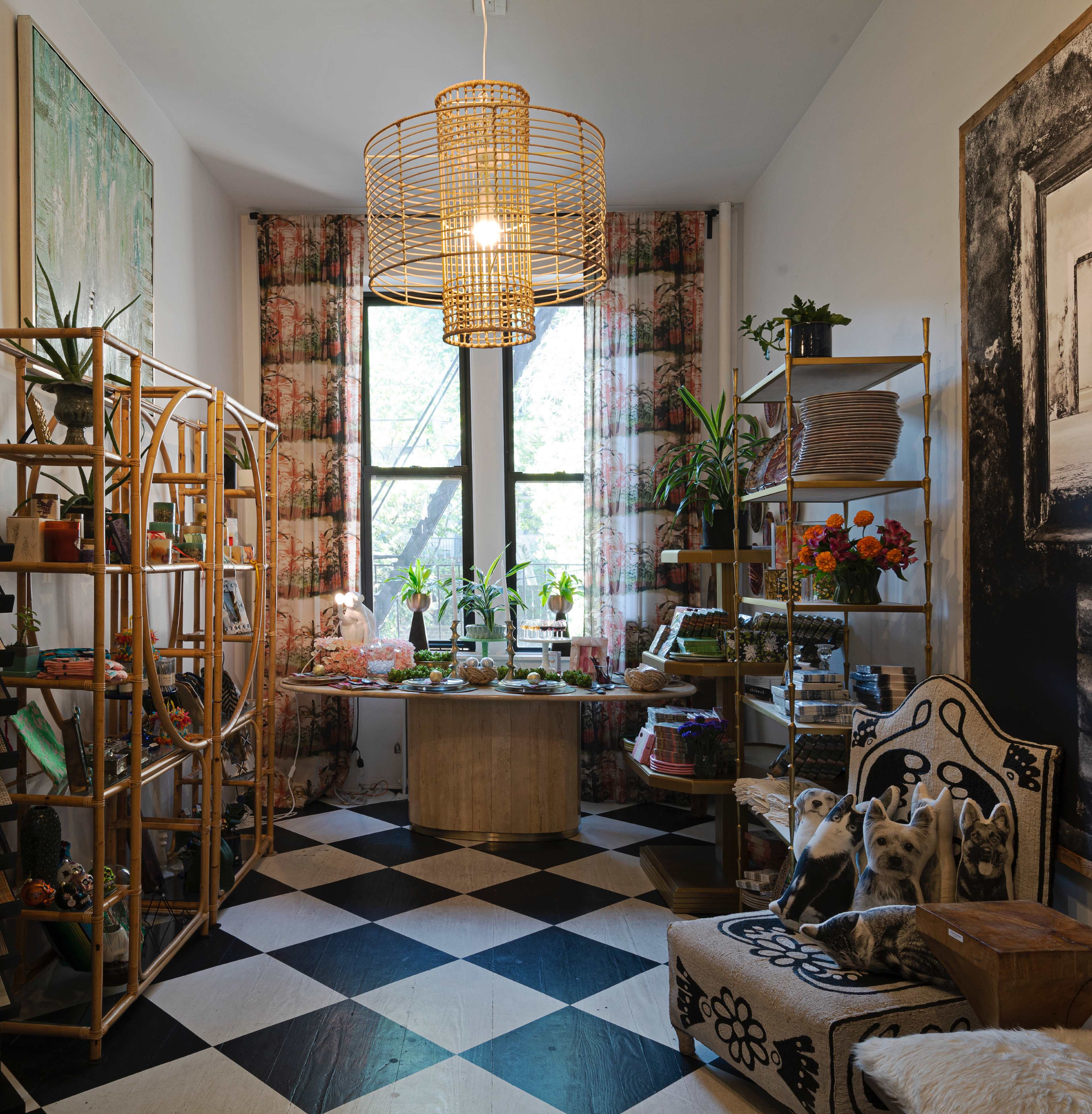 A well-furnished room featuring a round table surrounded by plants, shelves filled with various items, and a patterned chair, all set against a backdrop of large windows.