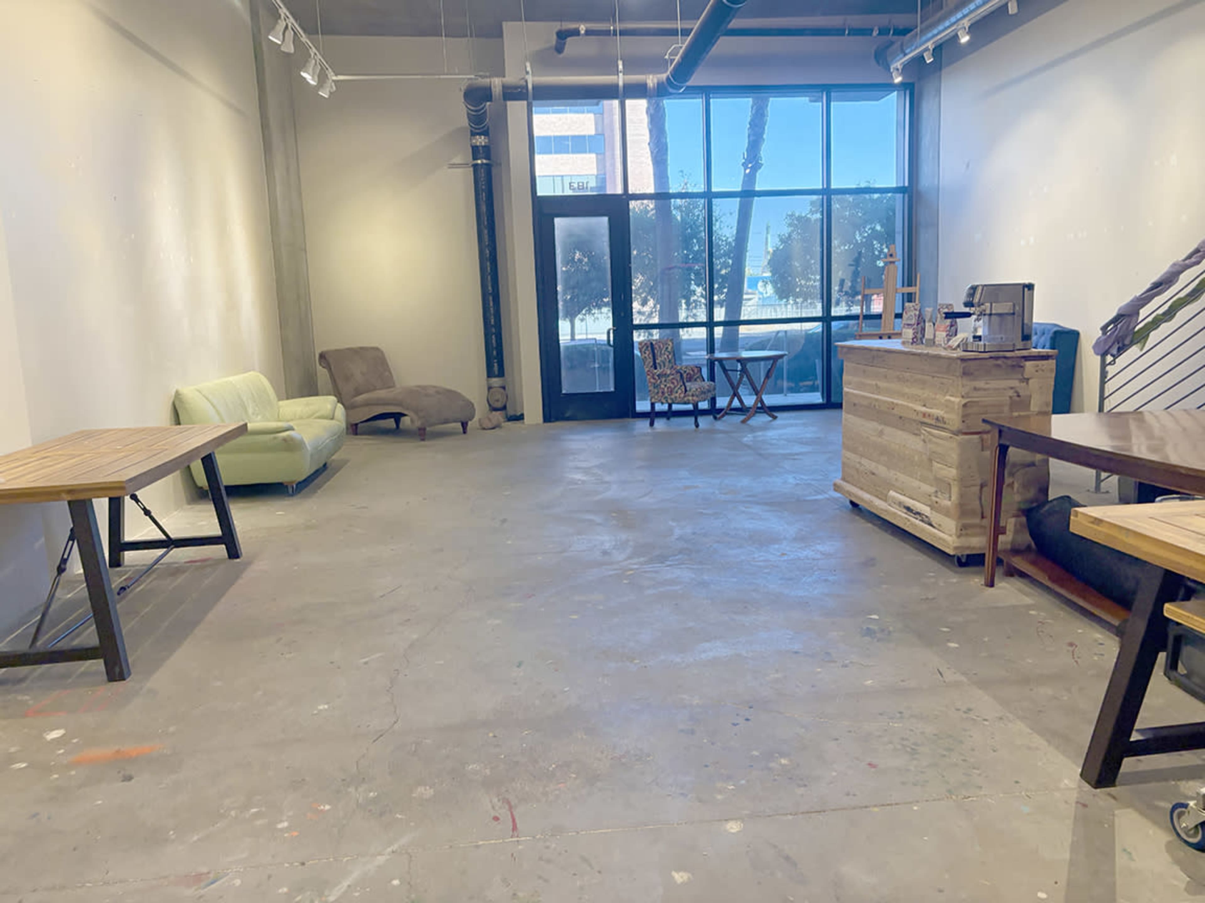 The image shows a spacious, empty room with concrete flooring, featuring two tables, a green couch, a brown chair, and large windows with natural light coming in.