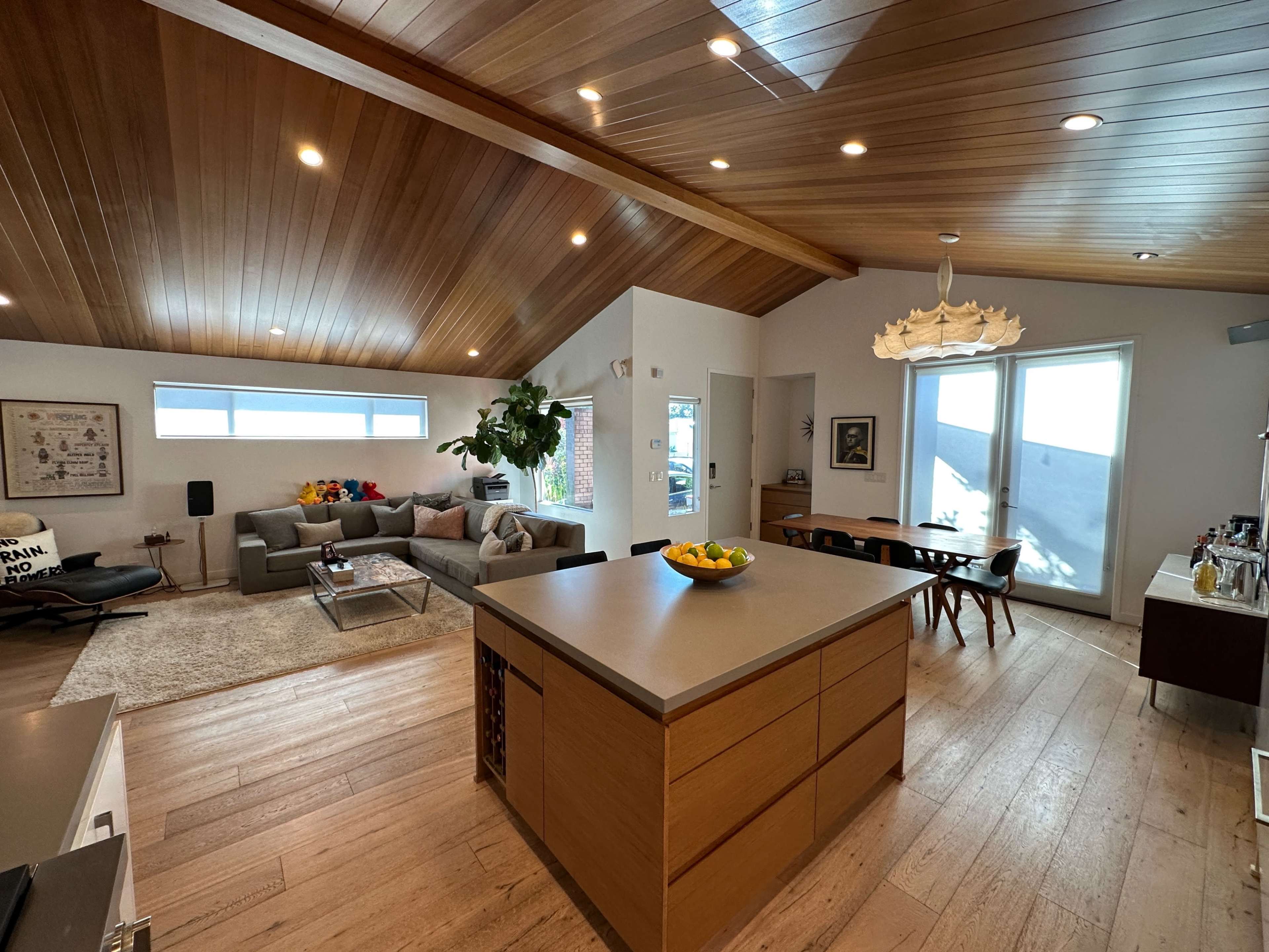 A modern open-concept living space with a wooden ceiling, a gray sofa, and a dining area featuring a table with a bowl of fruit.