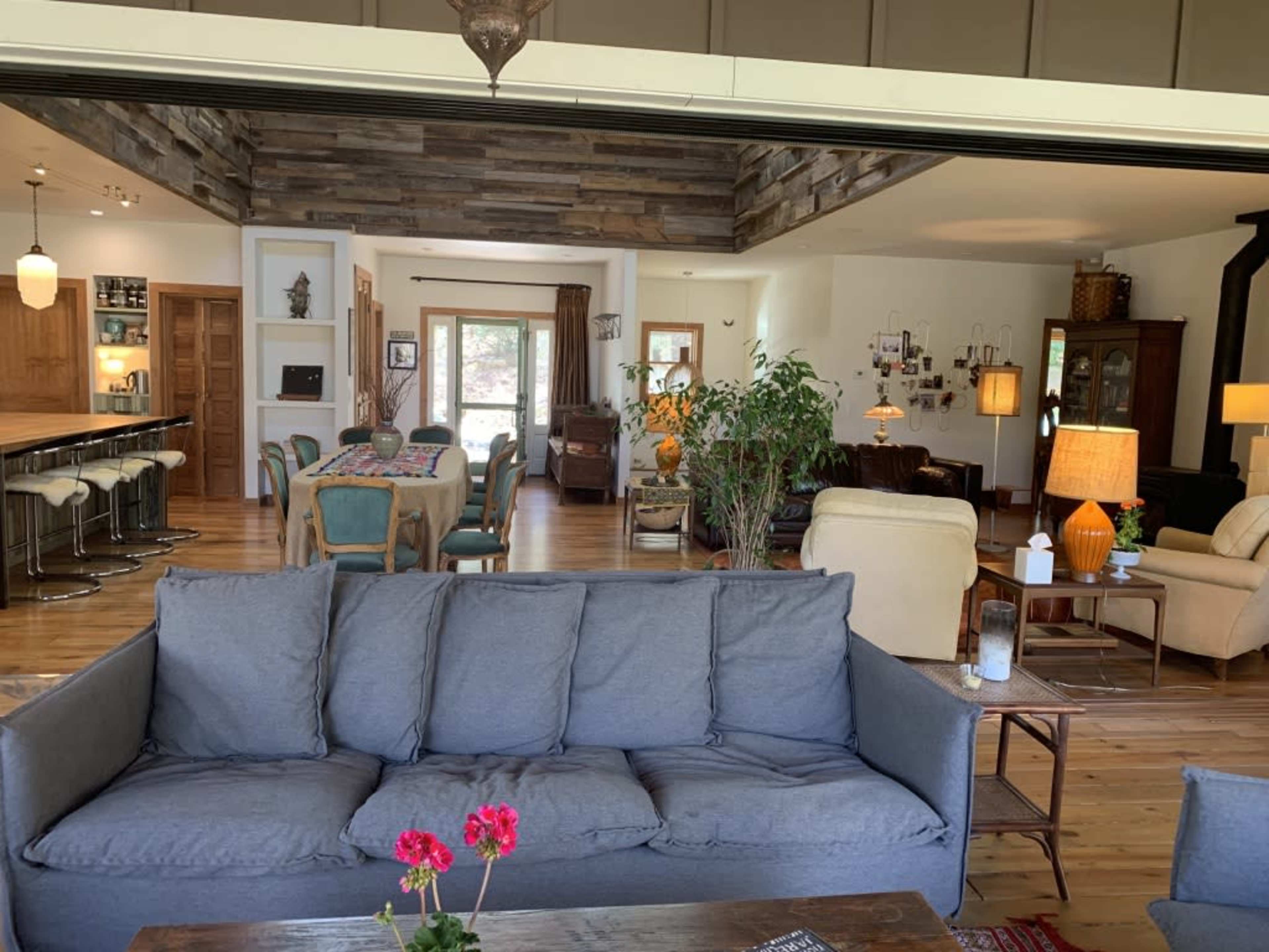 A spacious living area features a gray sofa with floral accents, wooden flooring, and a view of a dining space with a large table and various furnishings.