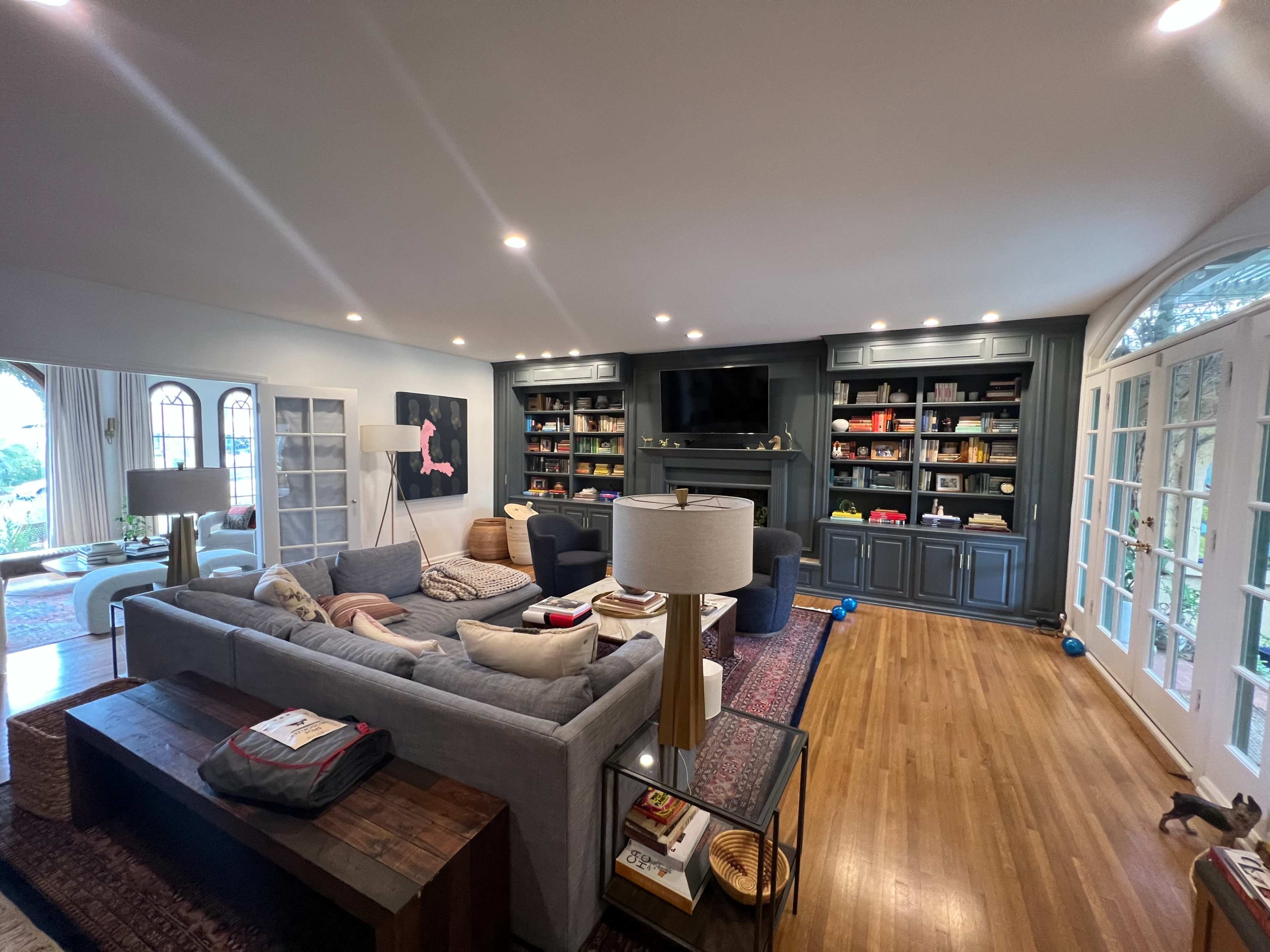A spacious living room with a gray sectional sofa, a wooden coffee table, built-in shelves filled with books and decor, and large windows allowing natural light.
