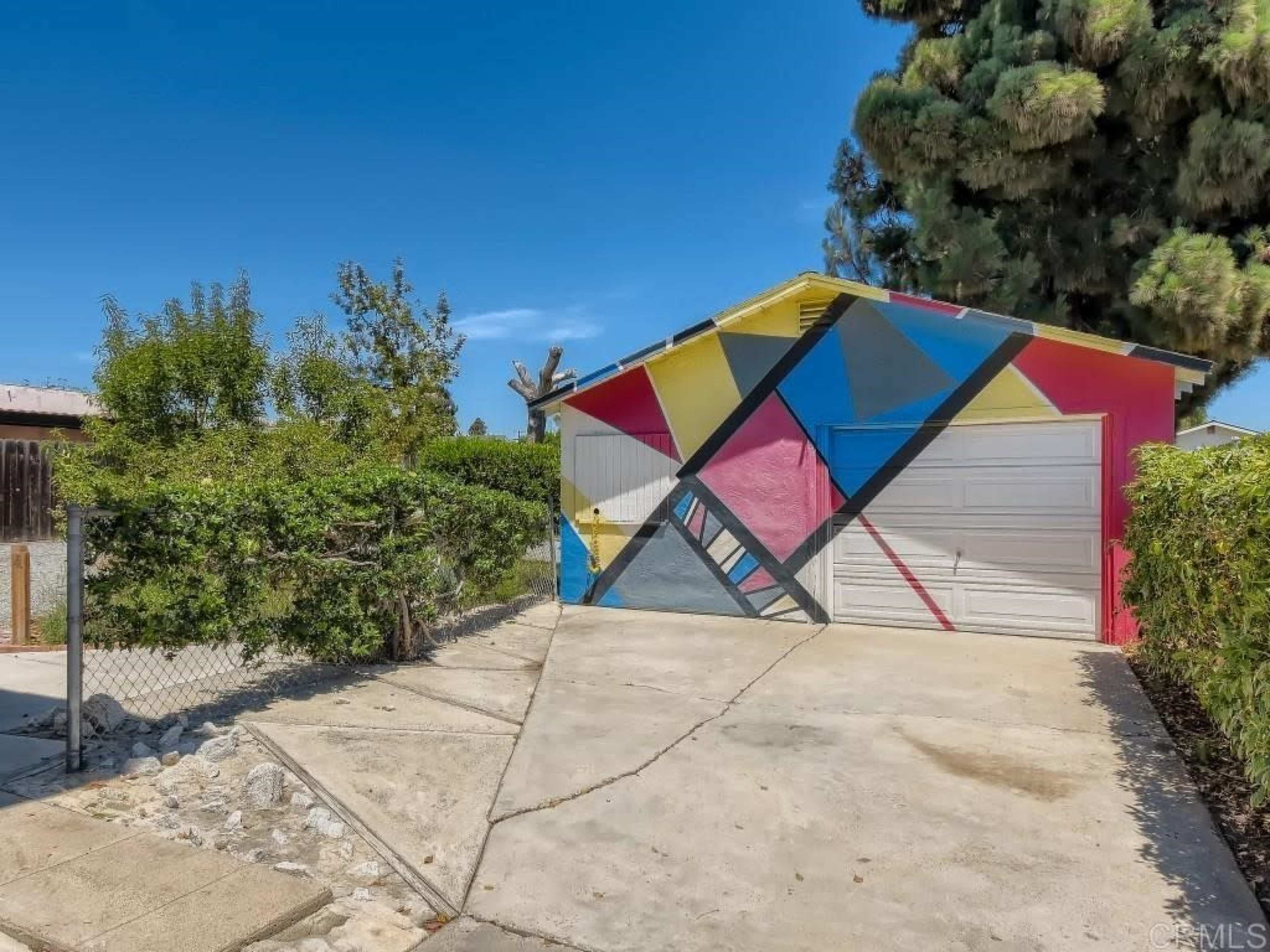 The image shows a colorful garage with geometric patterns on its exterior, situated next to a landscaped area and a pathway leading up to it.