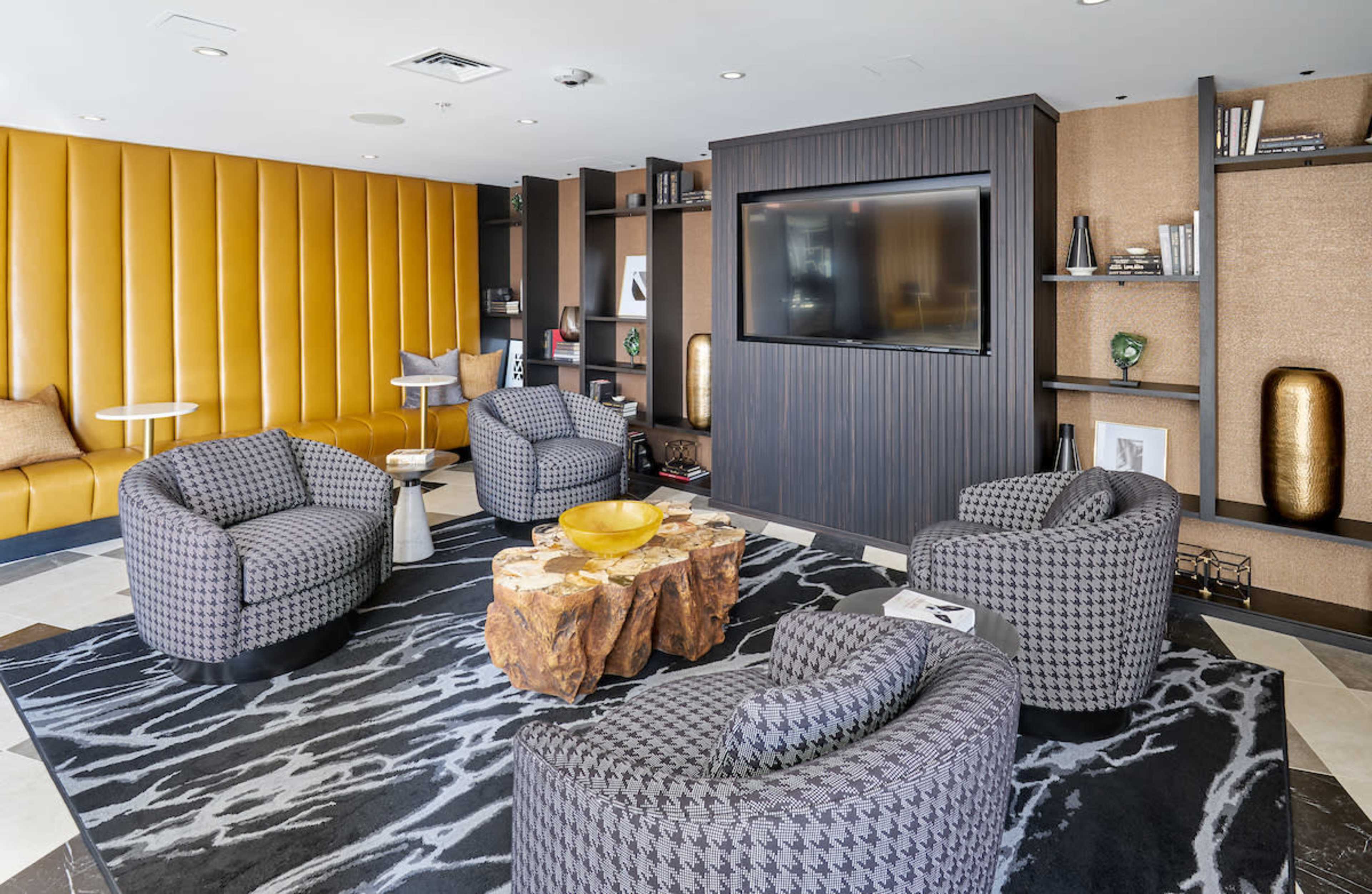 The image shows a modern lounge area with four patterned armchairs arranged around a wooden coffee table on a textured rug, with a television mounted on a dark wall.