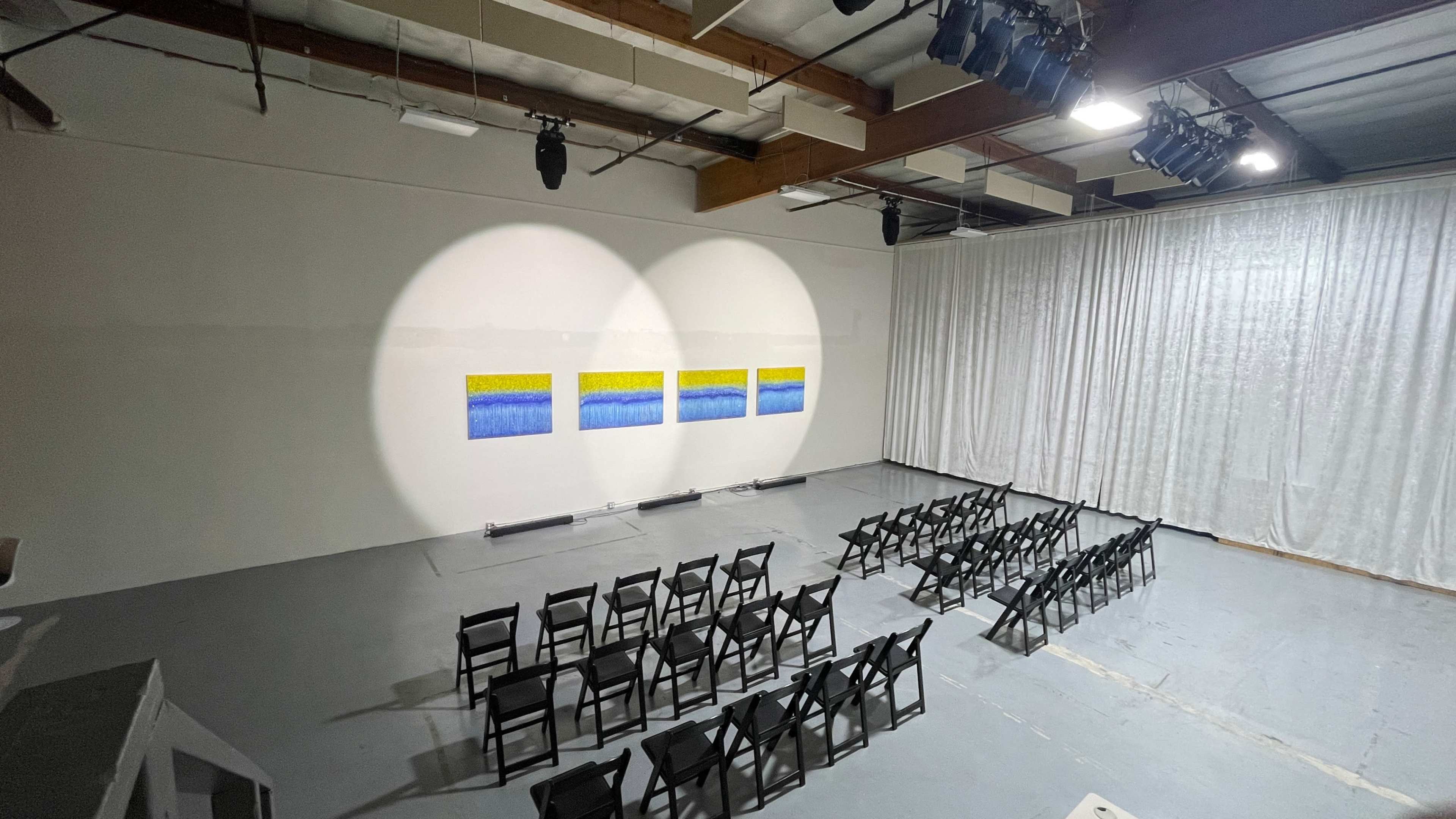 An art installation featuring a series of framed blue and yellow paintings is illuminated on a wall in a room set up with rows of black chairs.