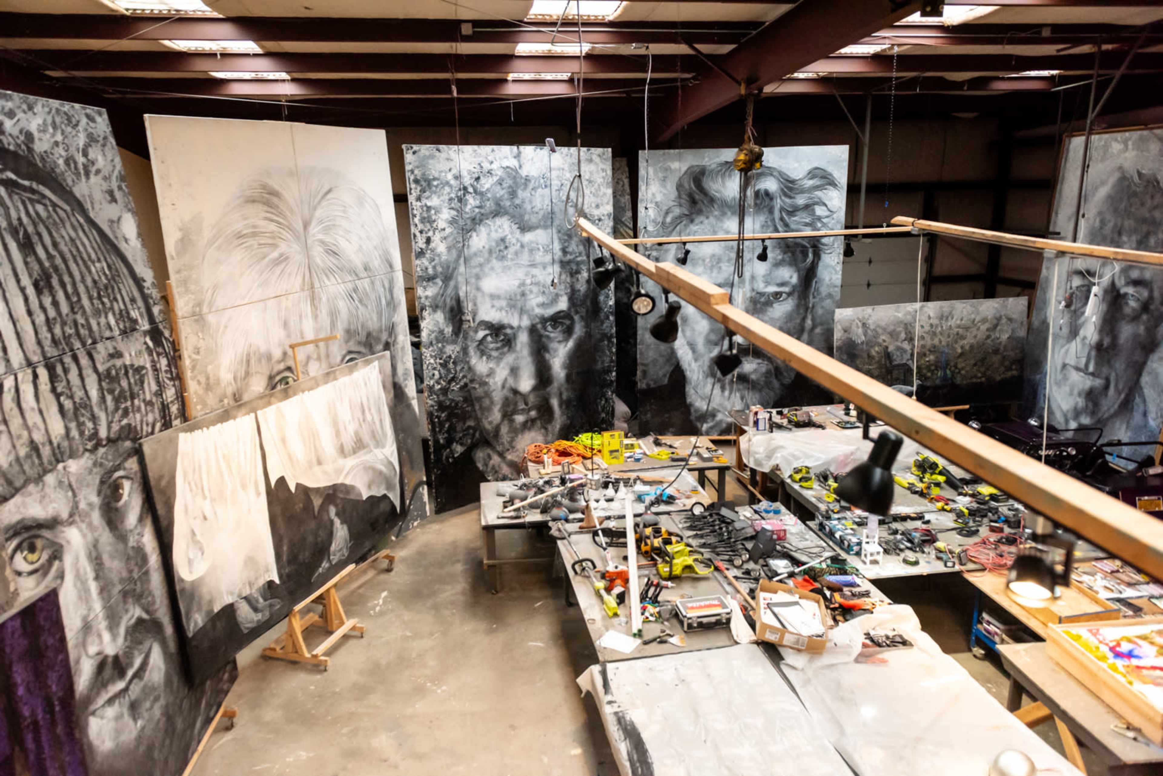 An artist's studio featuring large black-and-white portraits on the walls and a cluttered workspace with various art supplies and tools scattered on a table.