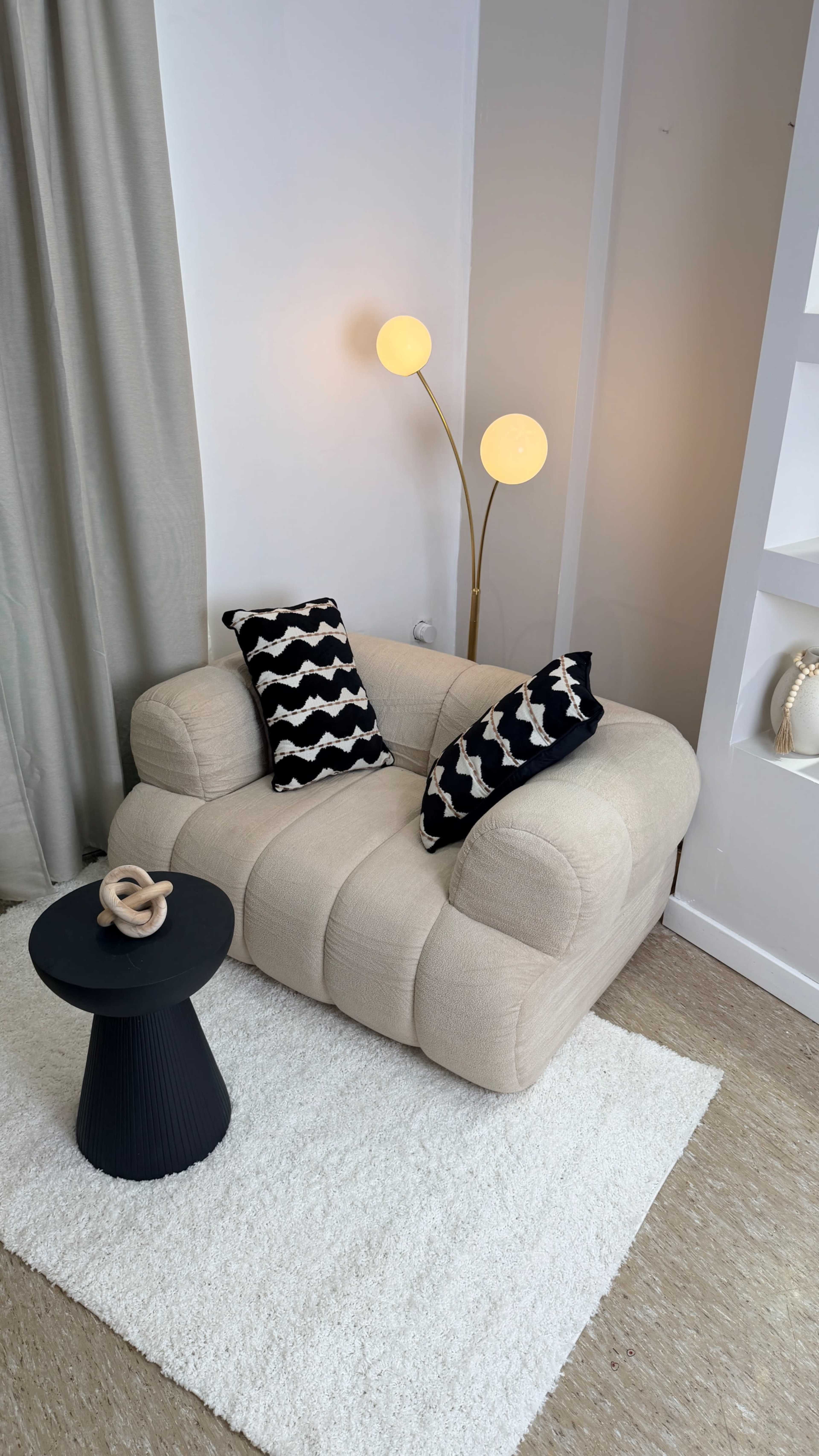 The image features a beige lounge chair with two patterned pillows, a small black side table, and a floor lamp with two globes in a softly lit room.