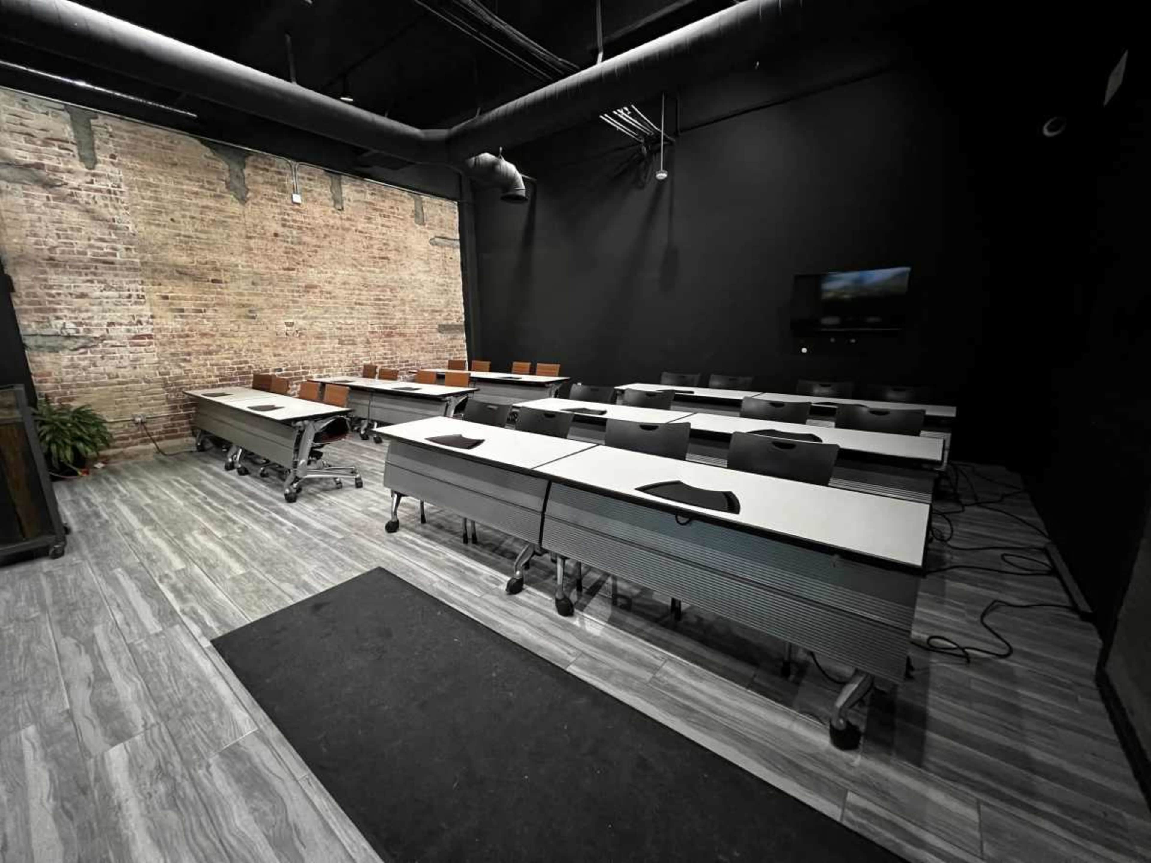 The image shows a modern classroom with movable tables and chairs arranged in rows against a textured brick wall.