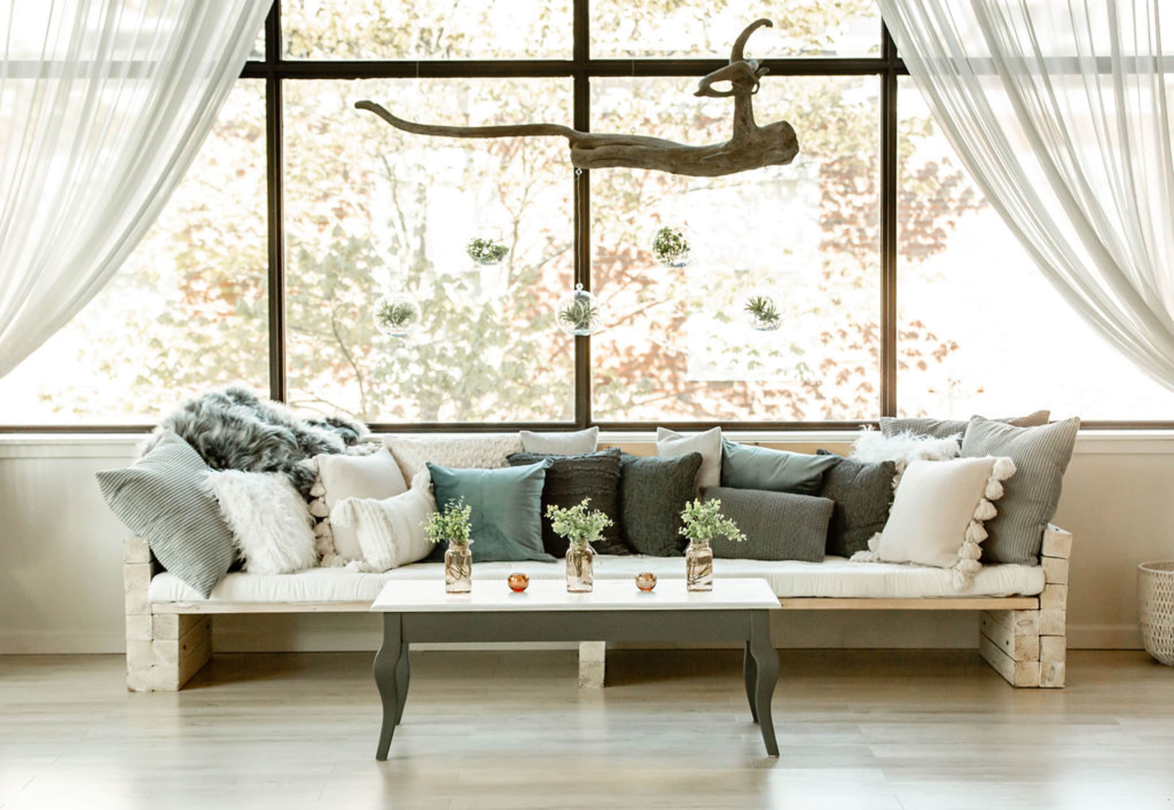 A minimalist sofa with multiple decorative pillows is positioned in front of a large window, adorned with sheer curtains and a few hanging plants.