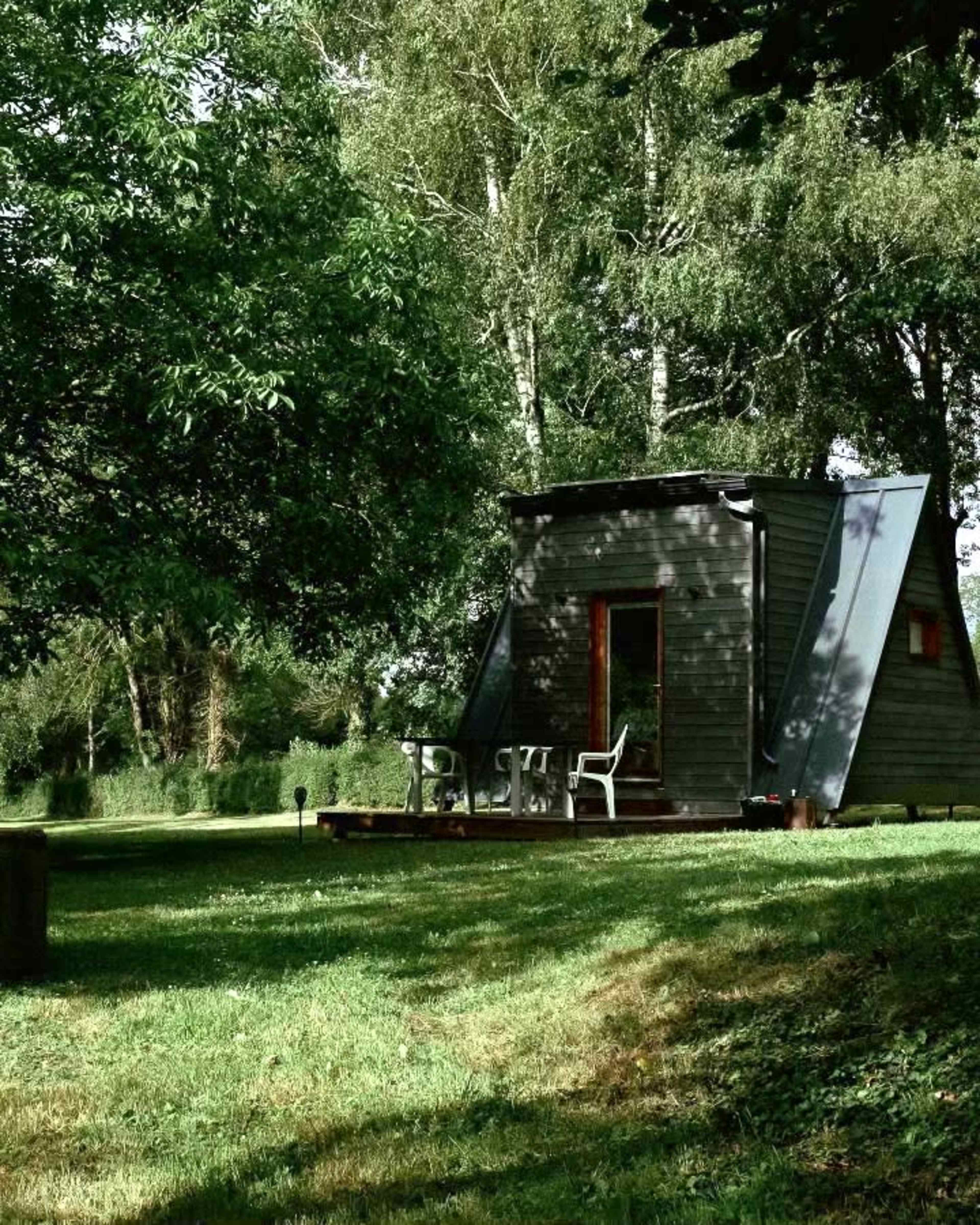 A small, modern cabin with an angled roof sits on a green lawn surrounded by trees.
