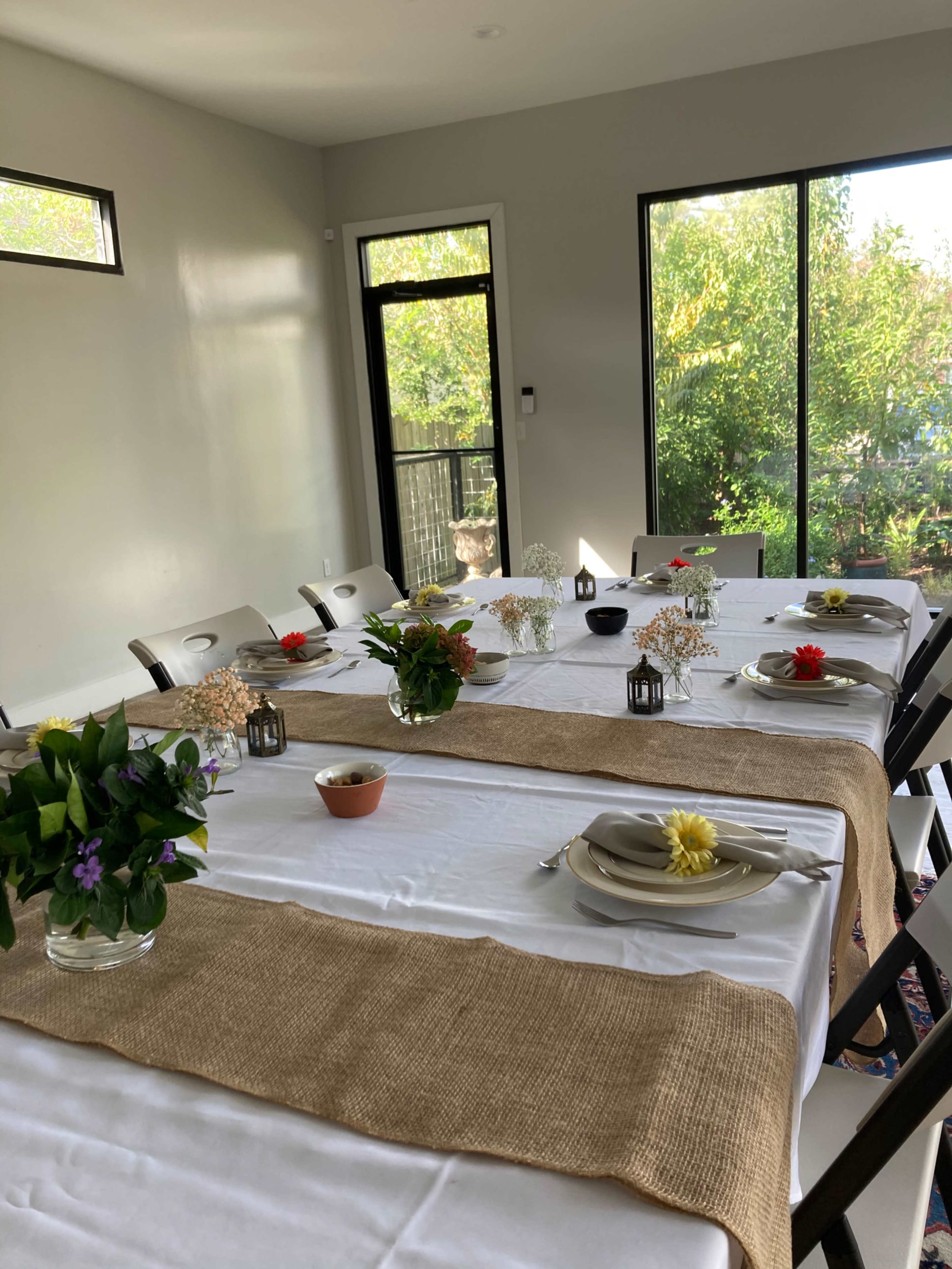 A dining room features two tables set with white tablecloths, decorated with small floral arrangements and simple dishware, overlooking a view of greenery through large windows.
