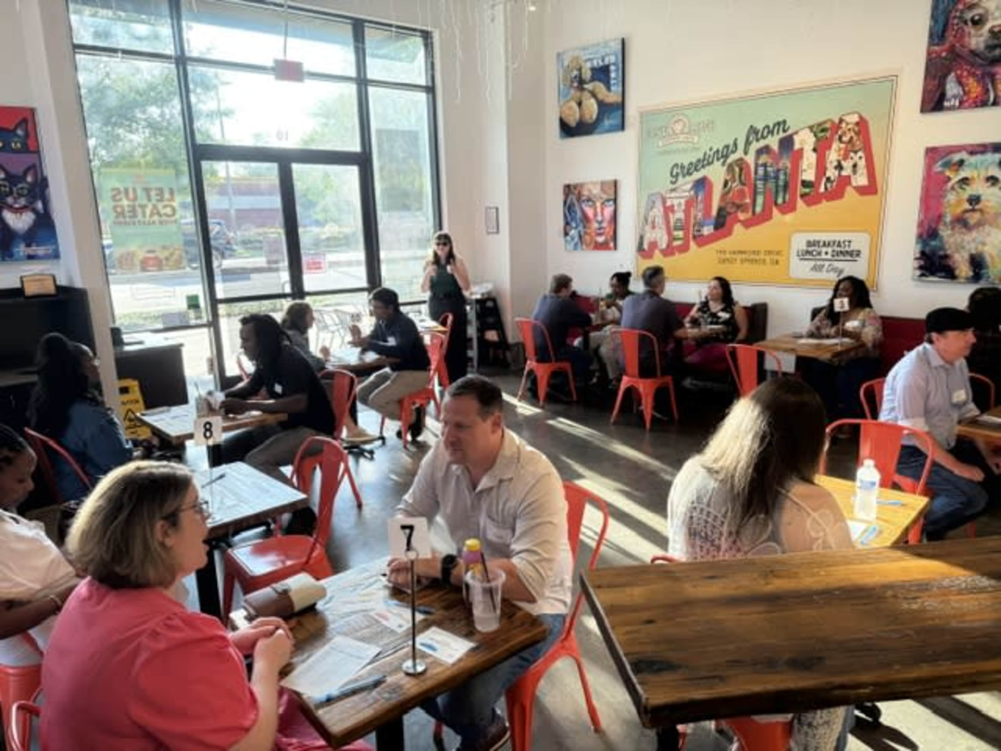 A busy café interior features people dining at wooden tables, with bright red chairs and colorful artwork on the walls.
