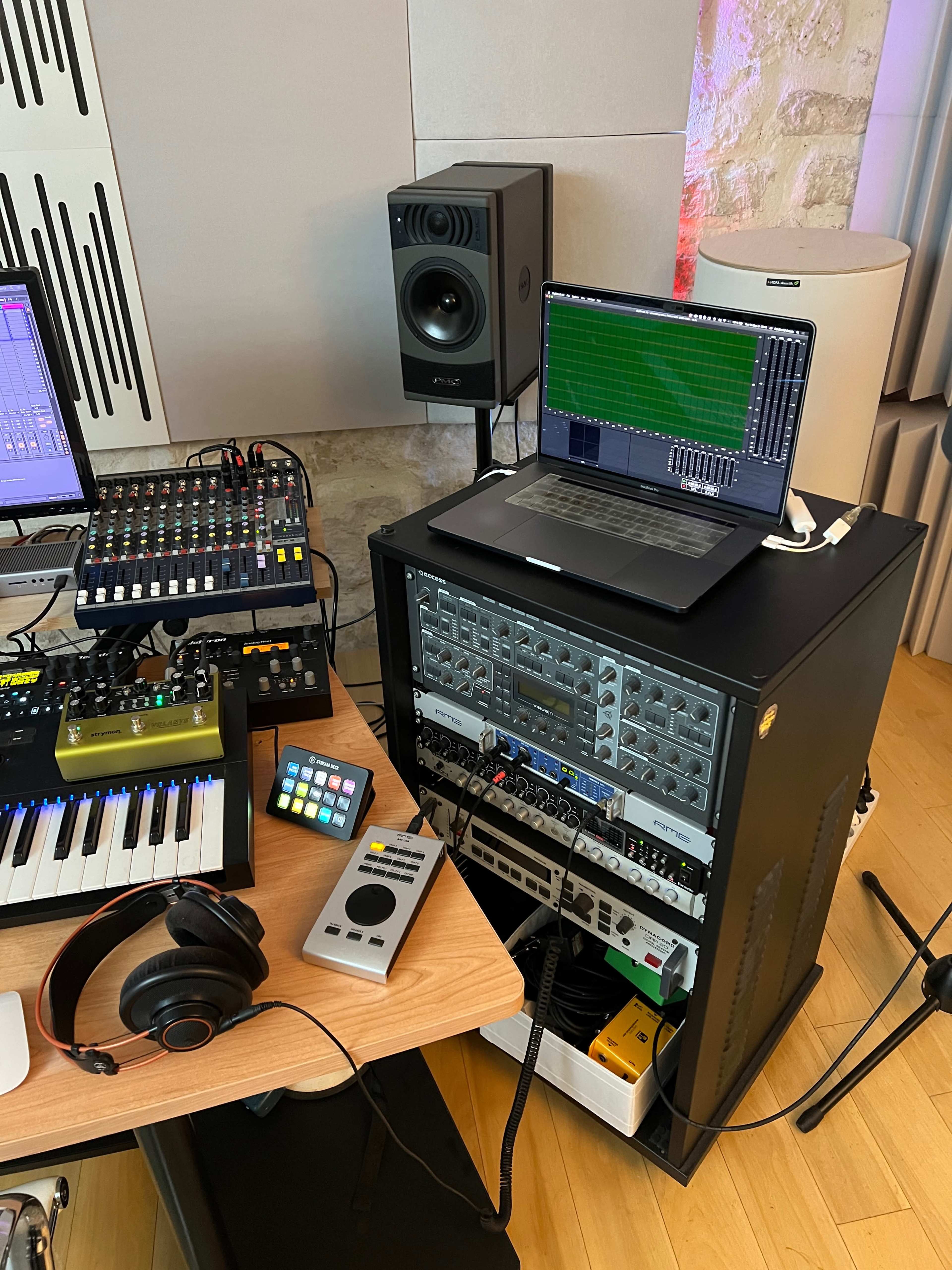 A music production workspace features a laptop, audio mixer, synthesizers, and studio monitors set on a wooden desk.