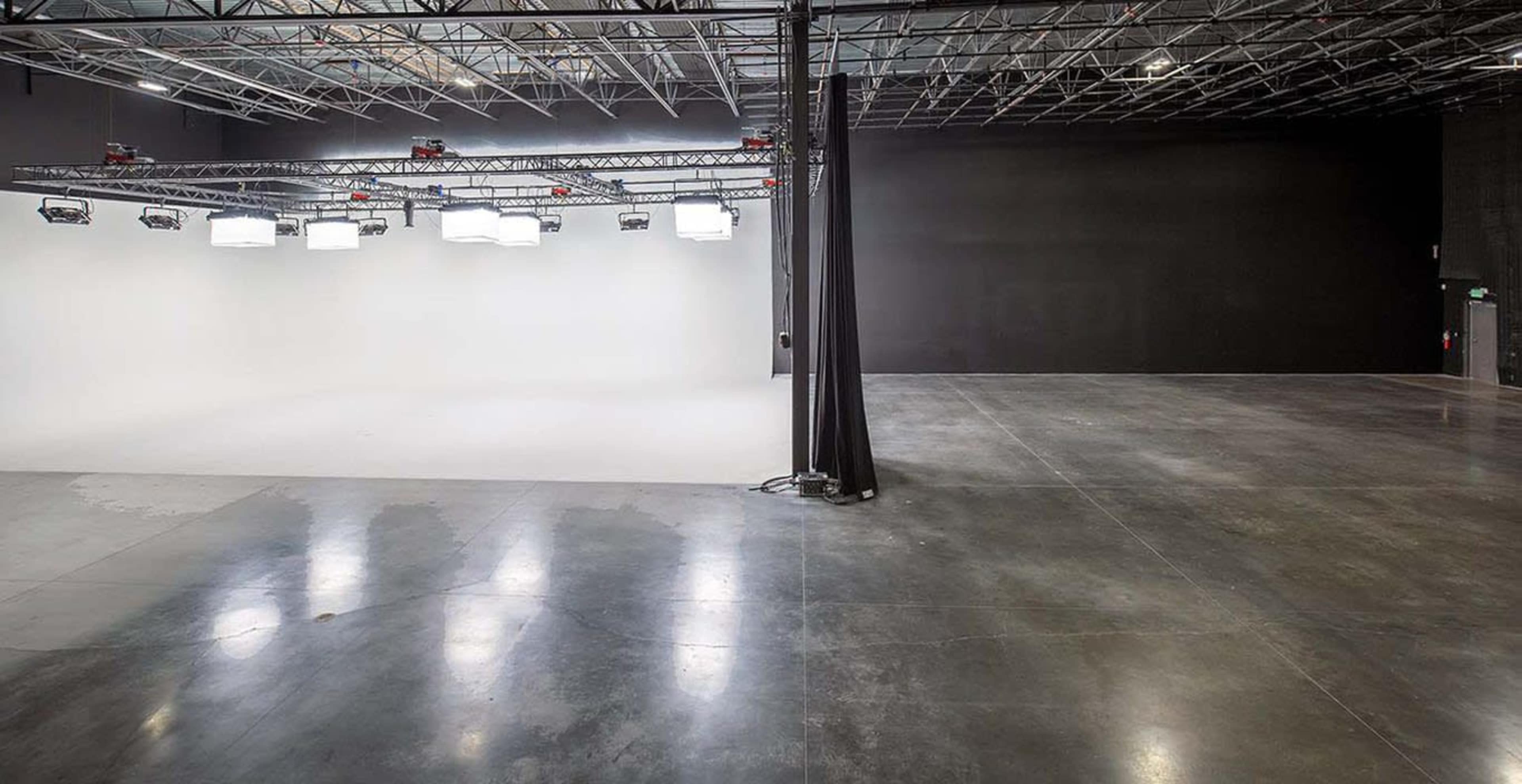 A large, empty studio space with a white backdrop on one side and a black wall on the other, featuring overhead lights and a polished concrete floor.