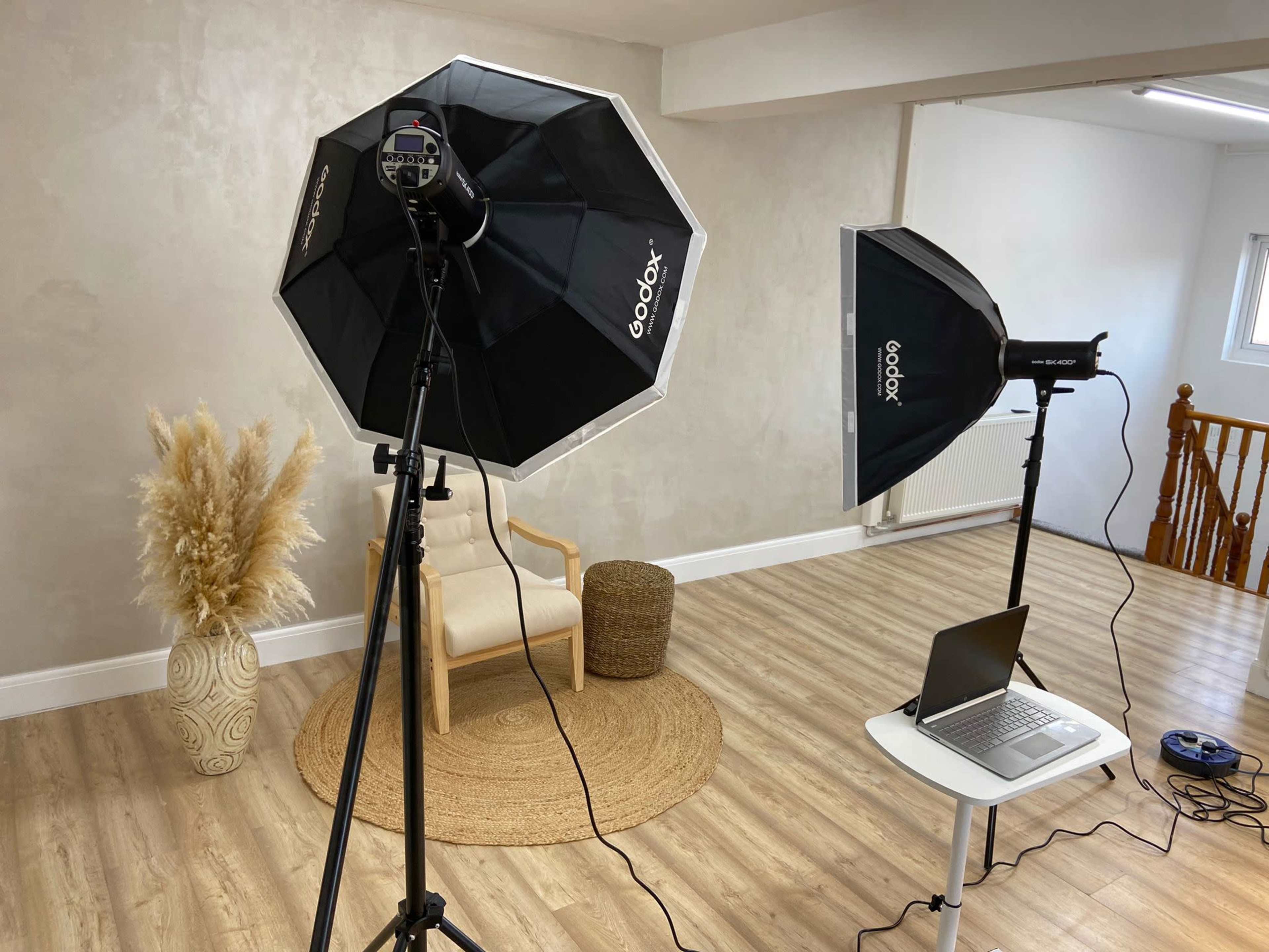 A well-lit studio setup featuring two softbox lights, a laptop on a small table, a chair, and a decorative plant in a corner on a wooden floor.