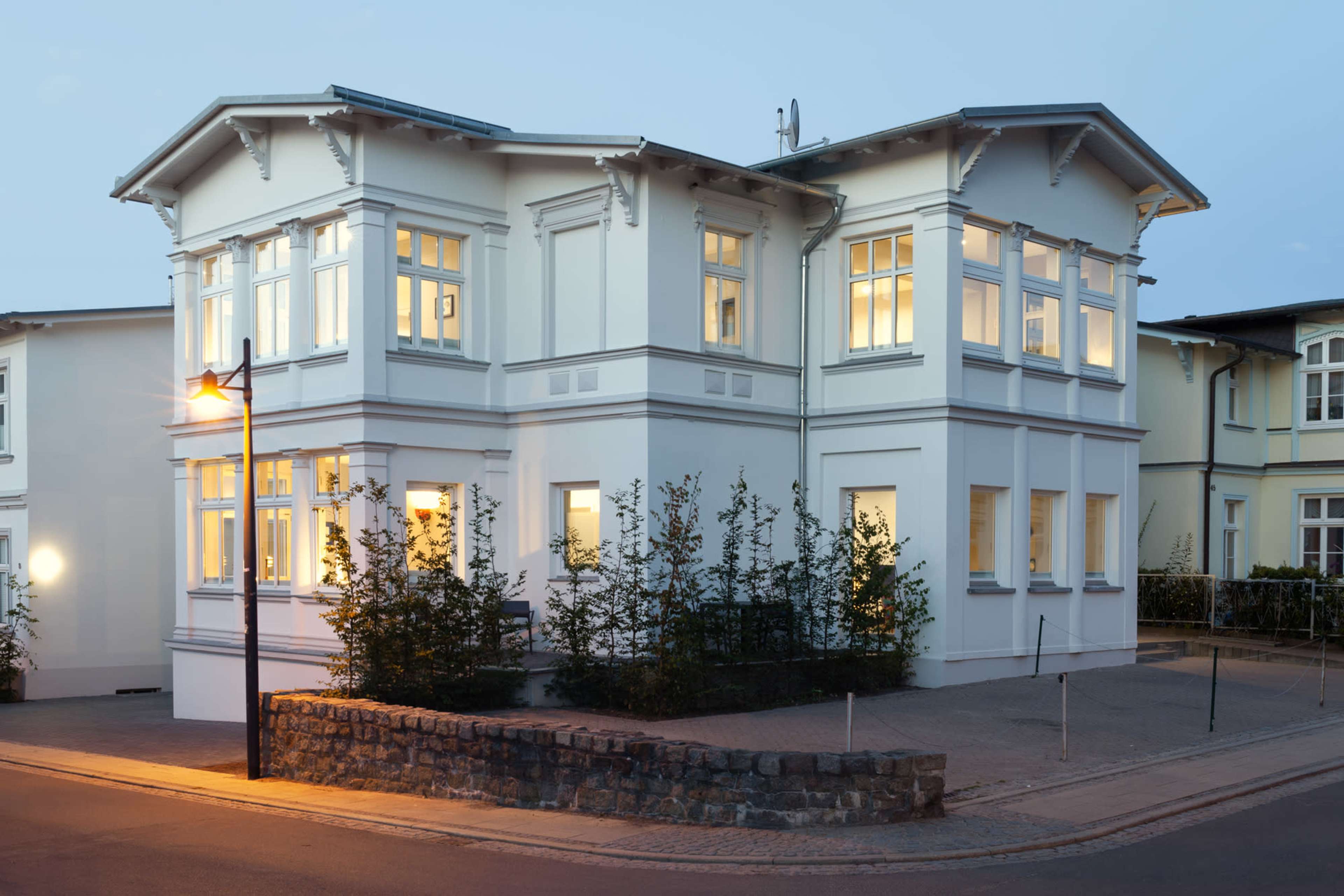 A two-story white house with large windows and a stone wall in front, situated at the corner of a street during twilight.