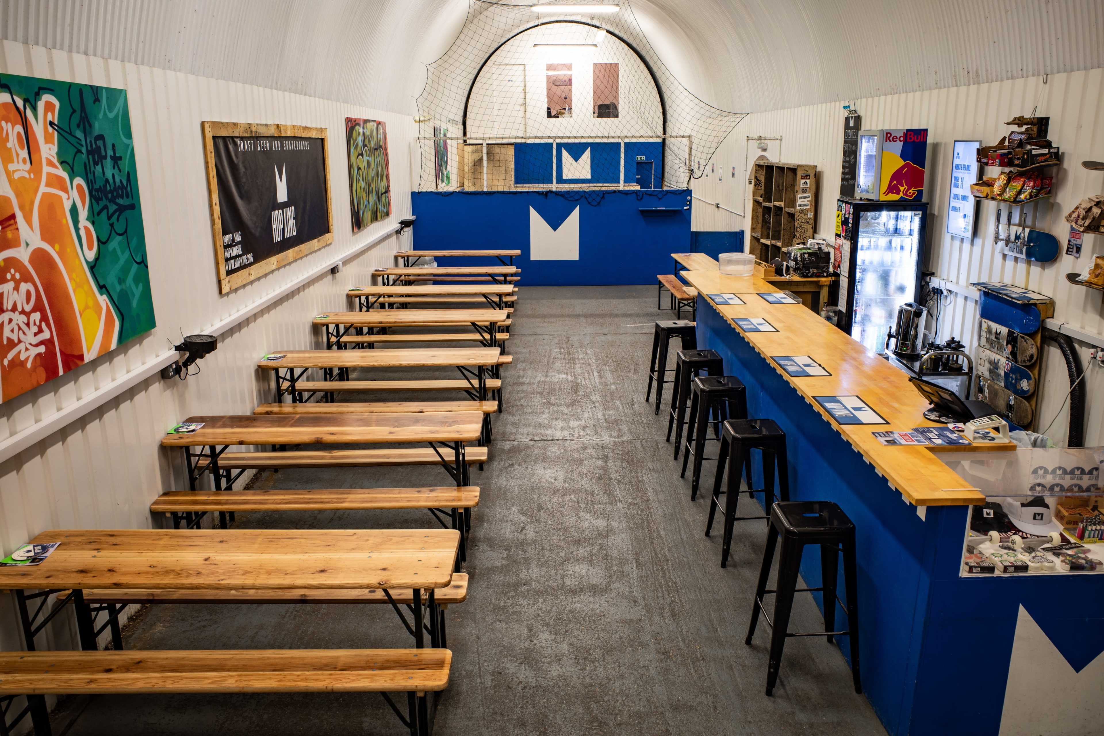 A spacious indoor venue with wooden tables, black stools, and a blue bar area, featuring graffiti on the walls and a netted section in the background.