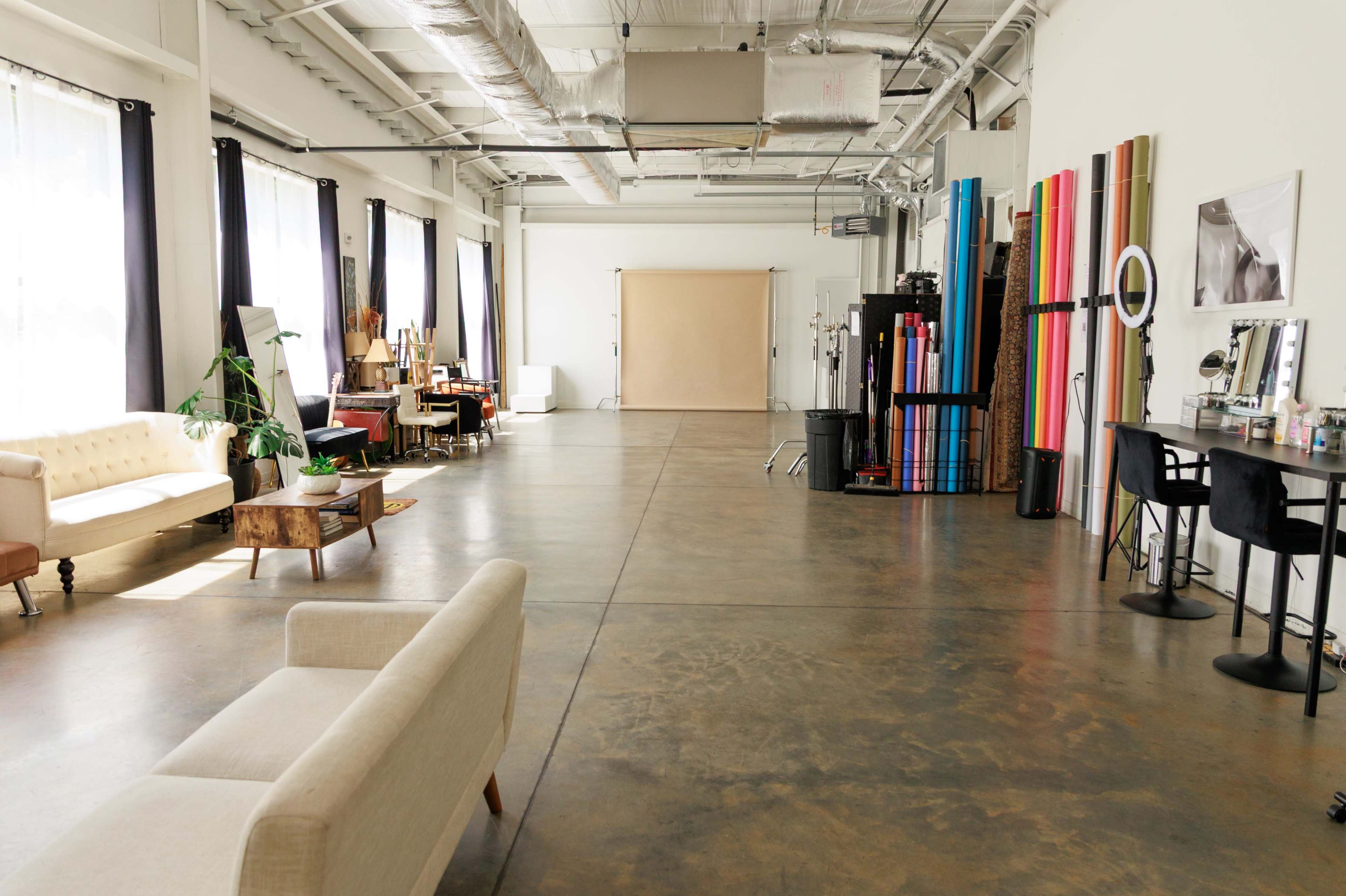 The image shows a spacious, well-lit studio featuring a mix of furniture, including a couch and tables, along with various backdrops arranged against the walls.