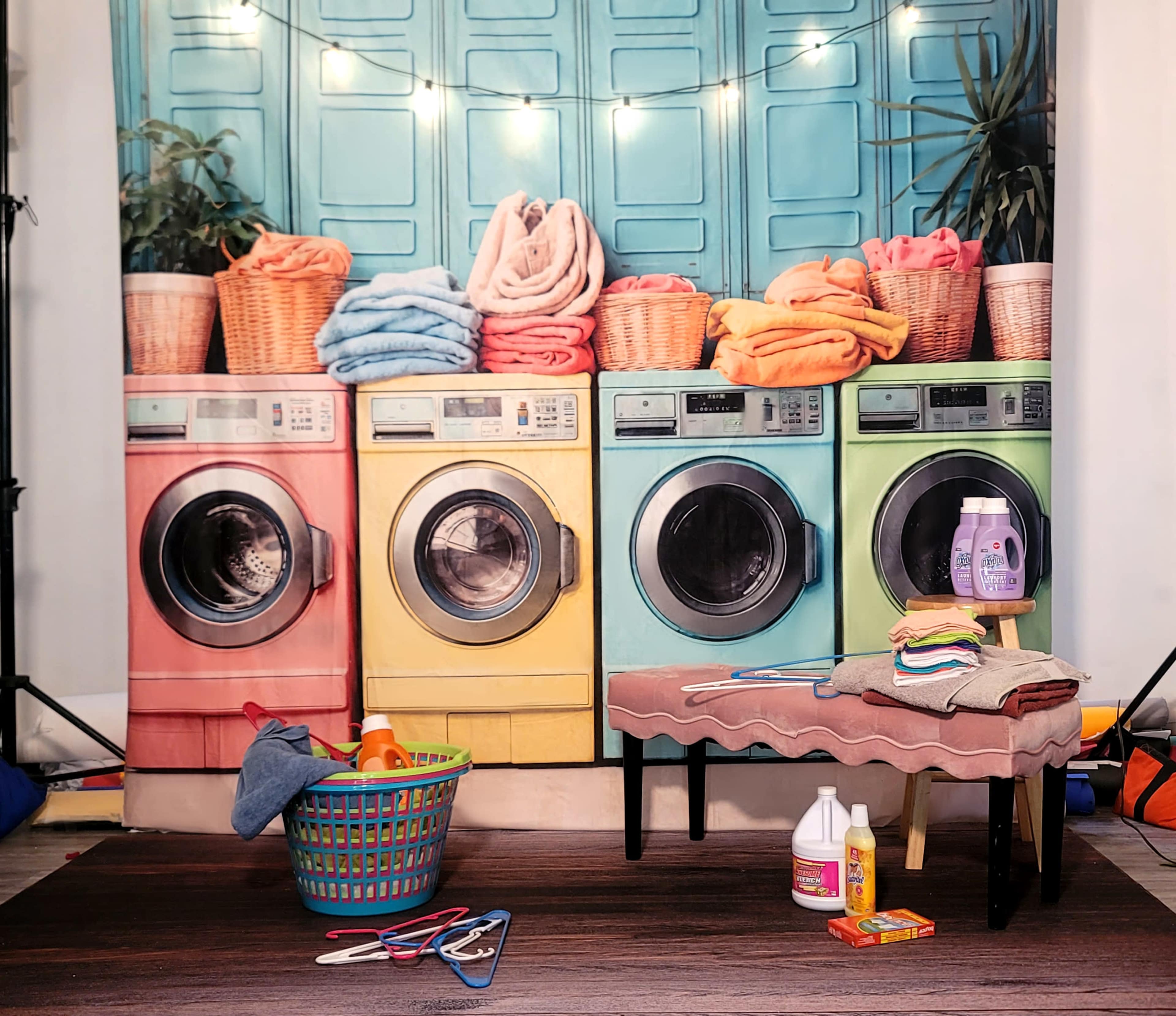The image features a vibrant laundry scene with three colorful washing machines, neatly stacked towels in baskets, and various laundry items arranged on a small bench.