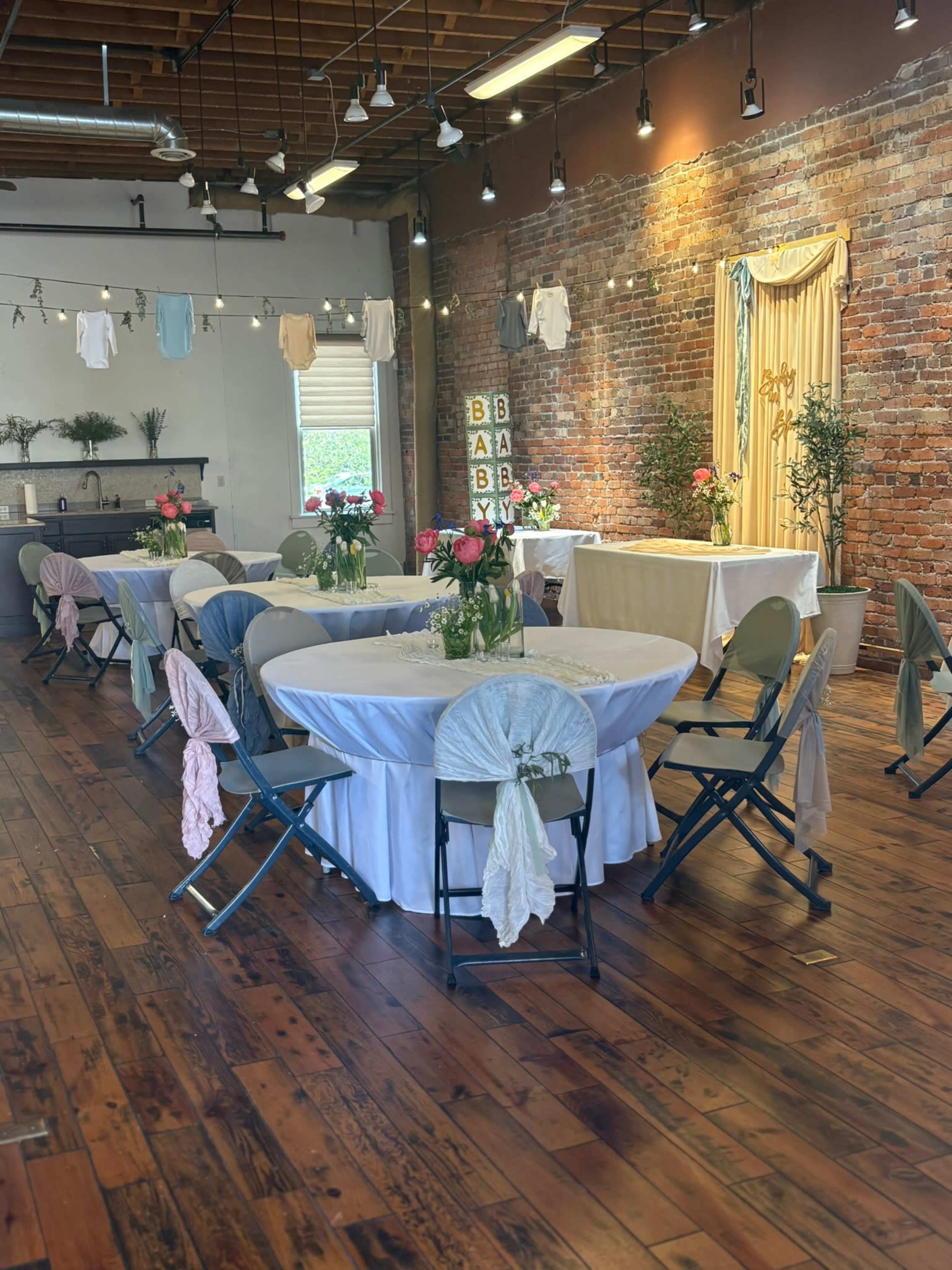 The image shows a spacious event venue with several round tables set with white tablecloths and floral centerpieces, decorated with draped fabric and hanging lights against a brick wall.